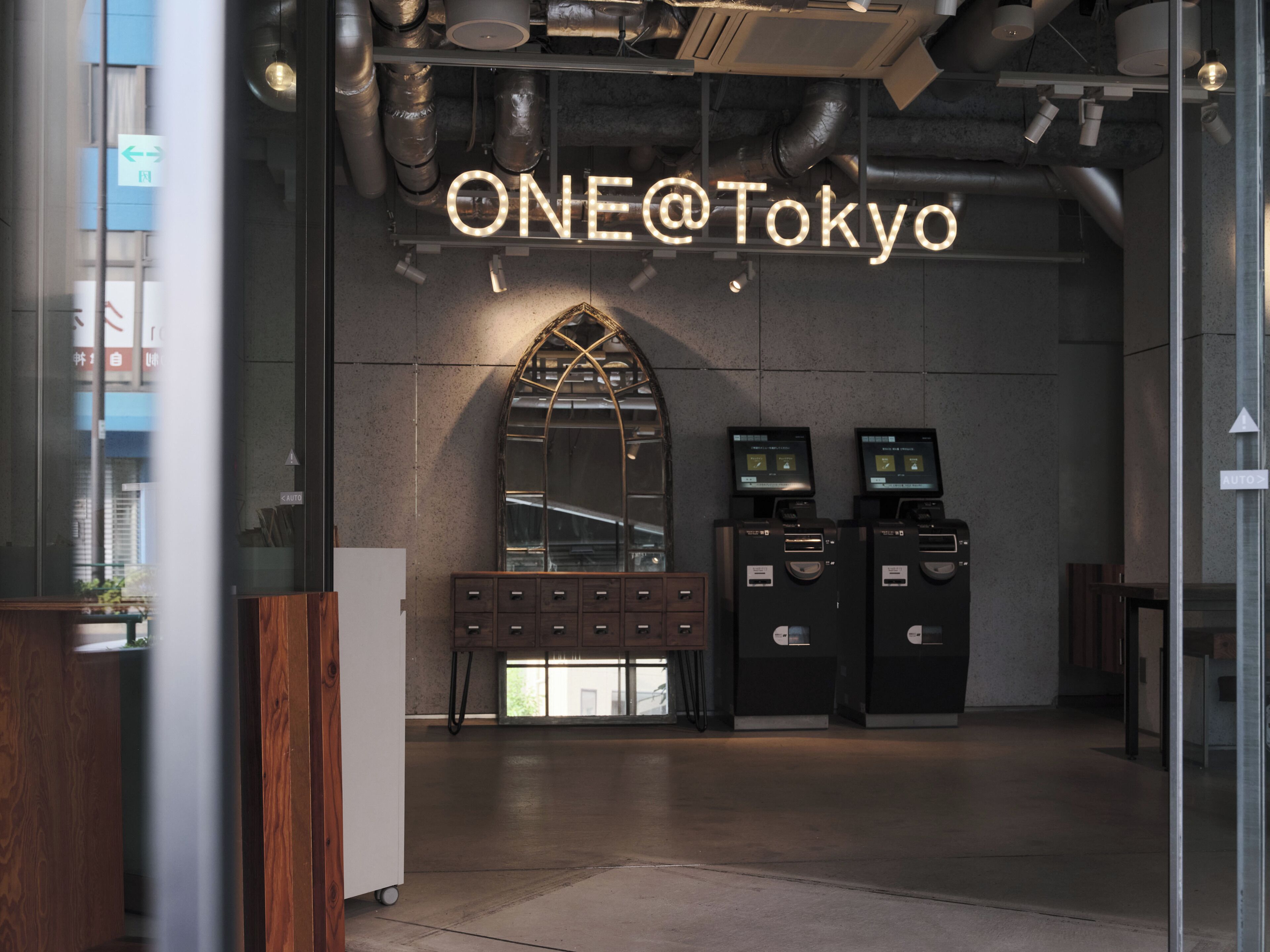 Ein modernes Interieur mit einem Neon-Schild mit der Aufschrift 'ONE@Tokyo', einem großen Bogenförmigen Spiegel und zwei Selbstbedienungs-Automaten.