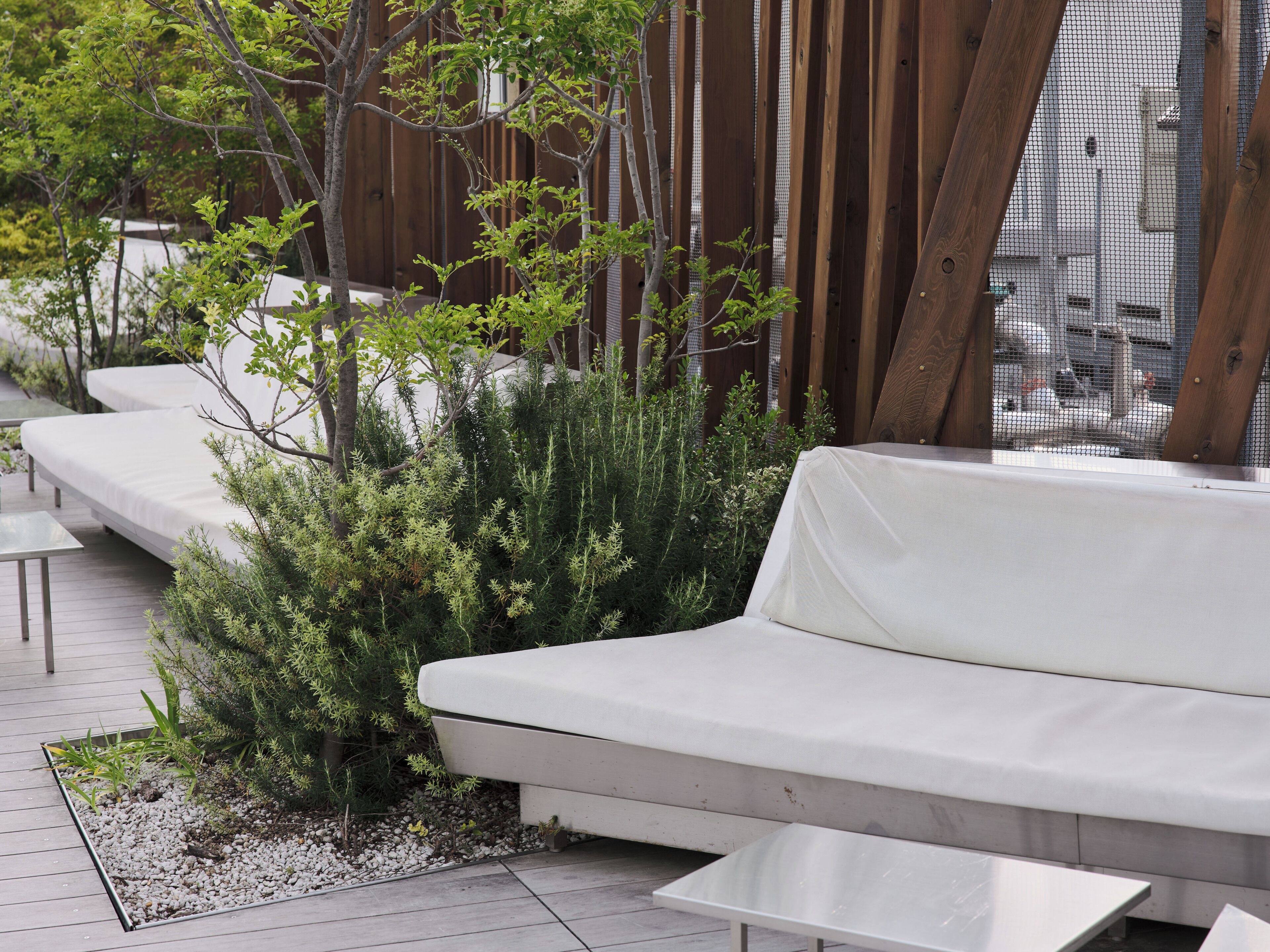 A modern outdoor seating area with white sofas and tables, surrounded by greenery and wooden structures.