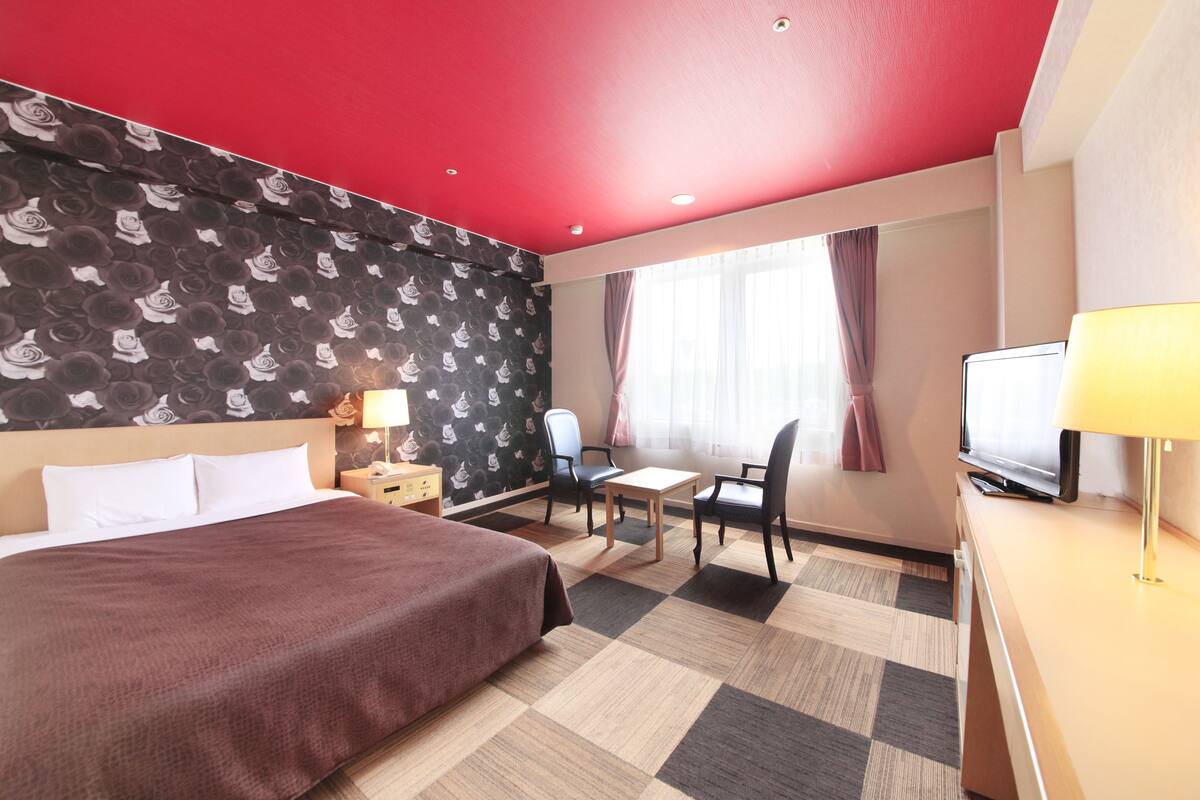 Habitación de hotel con cama, escritorio, silla, televisor y ventana con cortinas.