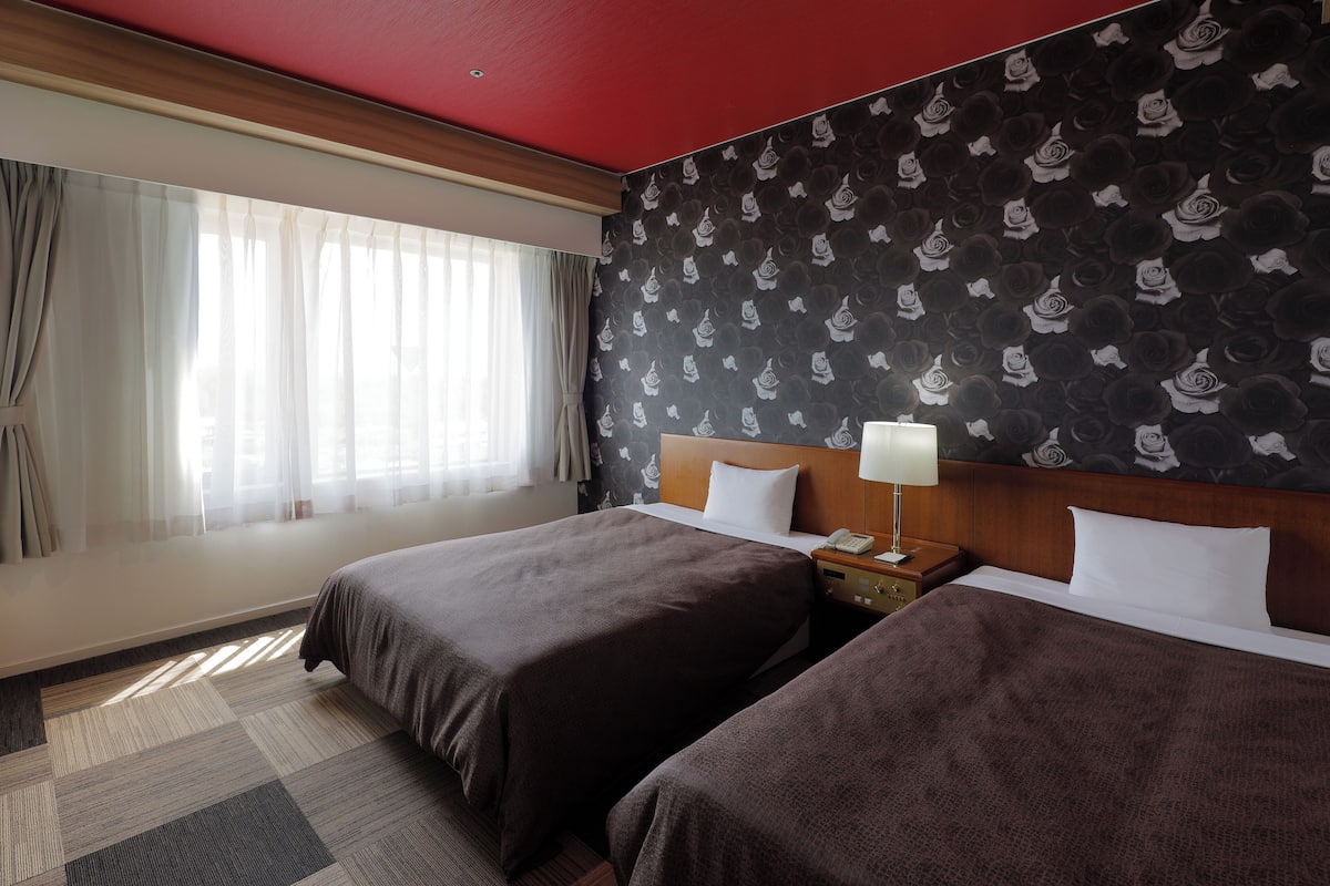 Habitación de hotel con dos camas, una mesita de noche con lámpara, pared estampada y ventana con cortinas translúcidas.