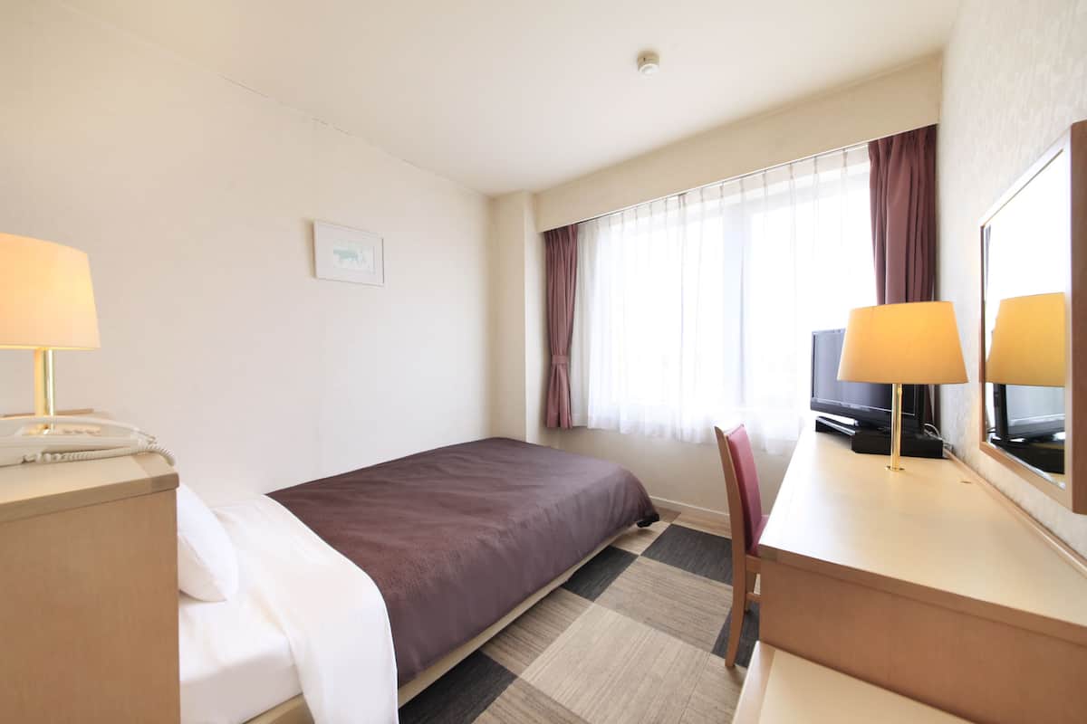 Una habitación de hotel con cama, escritorio con lámpara, silla, televisor y ventana con cortinas.