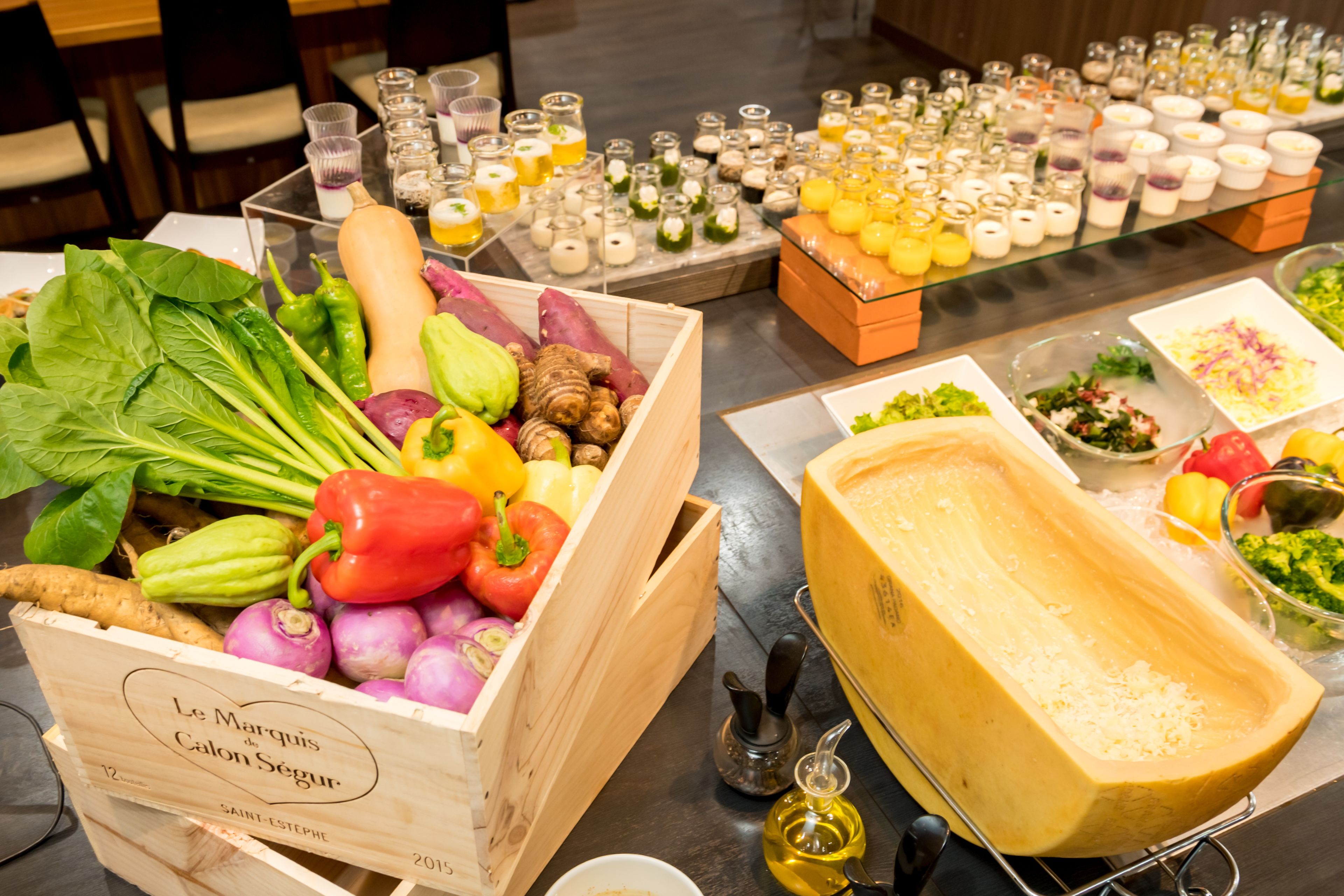 Une table de buffet proposant divers aliments, notamment une caisse en bois remplie de légumes, une planche de fromages et plusieurs petits pots de condiments.
