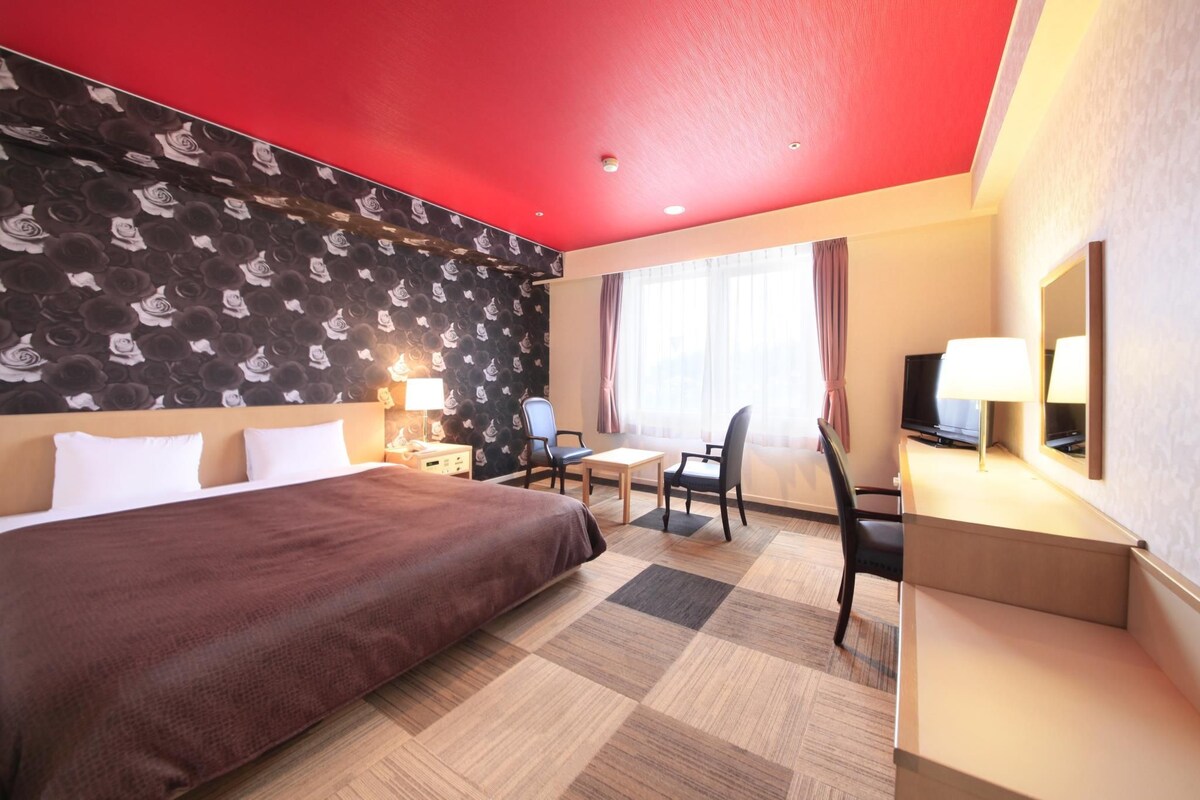 Habitación de hotel con una cama grande, un escritorio, una silla, un televisor y una ventana con cortinas.