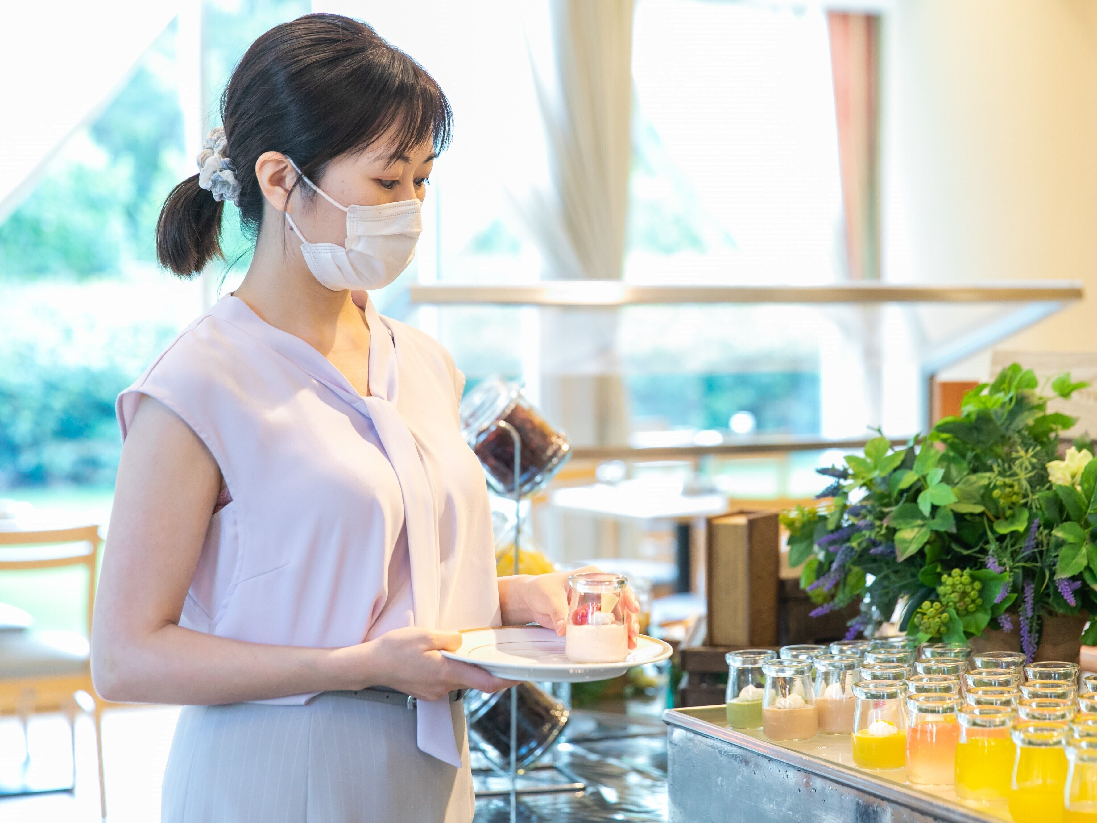Une femme tenant une assiette avec une boisson, debout dans une pièce avec une grande fenêtre et une plante en arrière-plan.