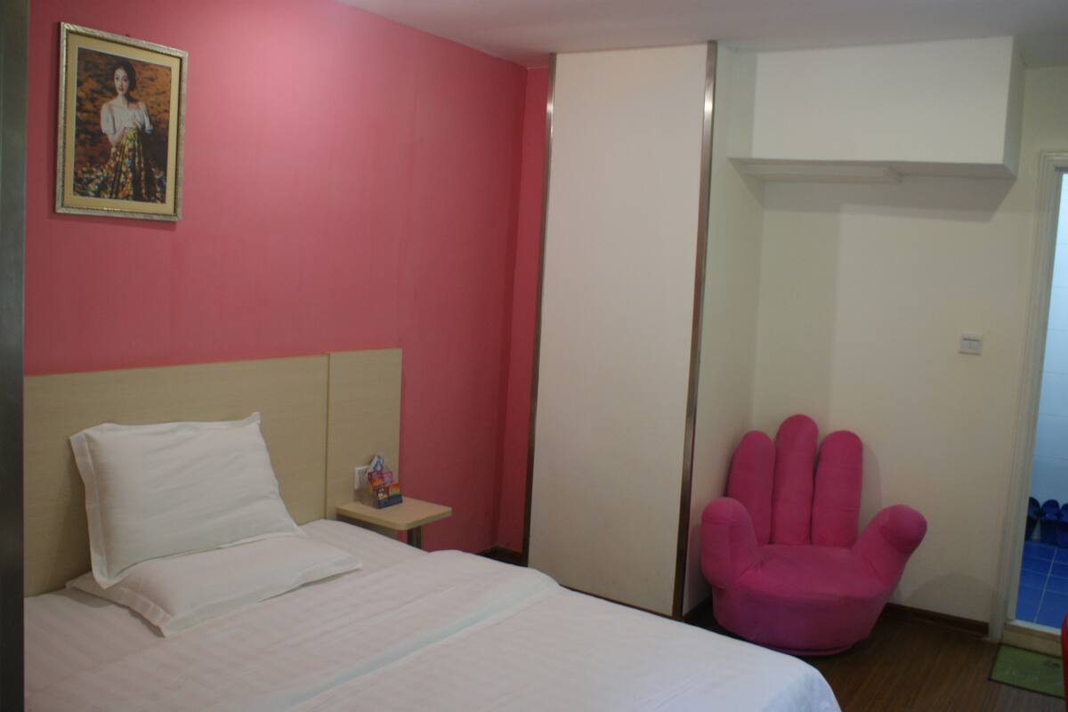 Um quarto com cama, mesa de cabeceira, uma cadeira rosa e um guarda-roupa.