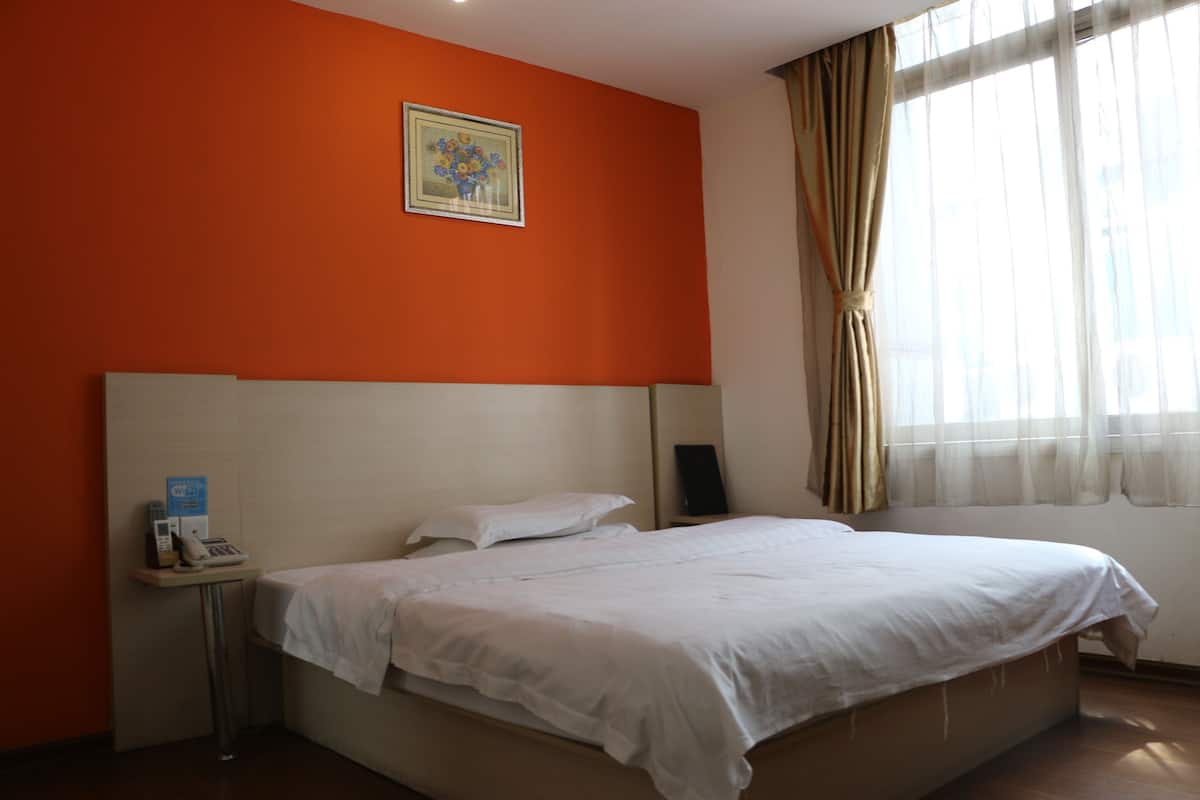 Quarto de hotel com parede laranja, cama com lençóis brancos, mesa de cabeceira com telefone e uma caixa, e janela com cortinas translúcidas.