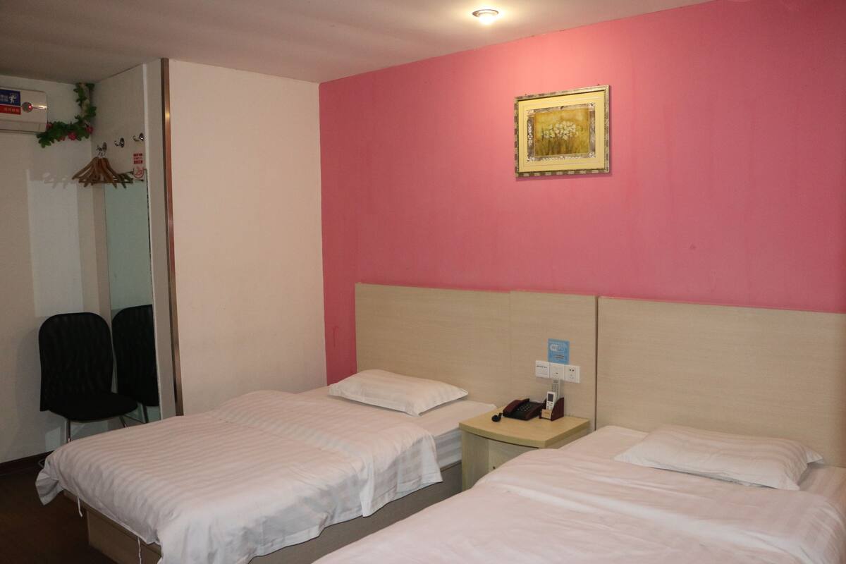 Quarto de hotel com duas camas de solteiro, uma pequena mesa de cabeceira com telefone, uma cadeira e um quadro na parede.