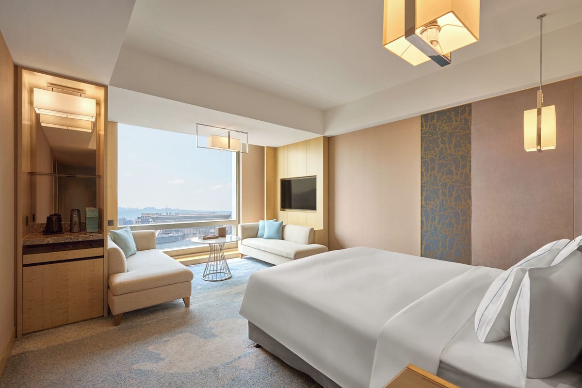 Quarto de hotel com cama, sofá, escrivaninha com espelho, televisão e uma grande janela com vista para a cidade.