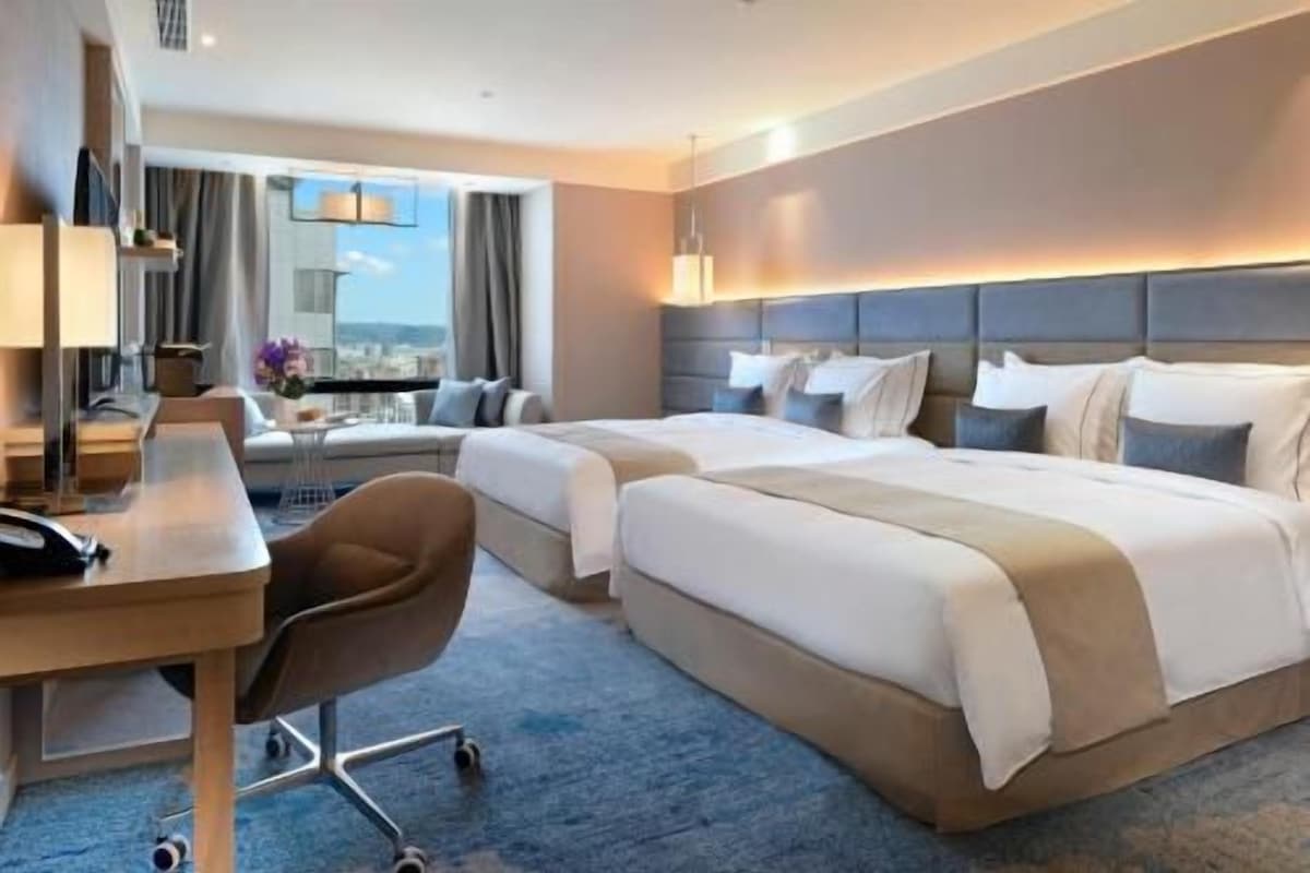 Habitación de hotel con dos camas, un escritorio, una silla y un ventanal grande.