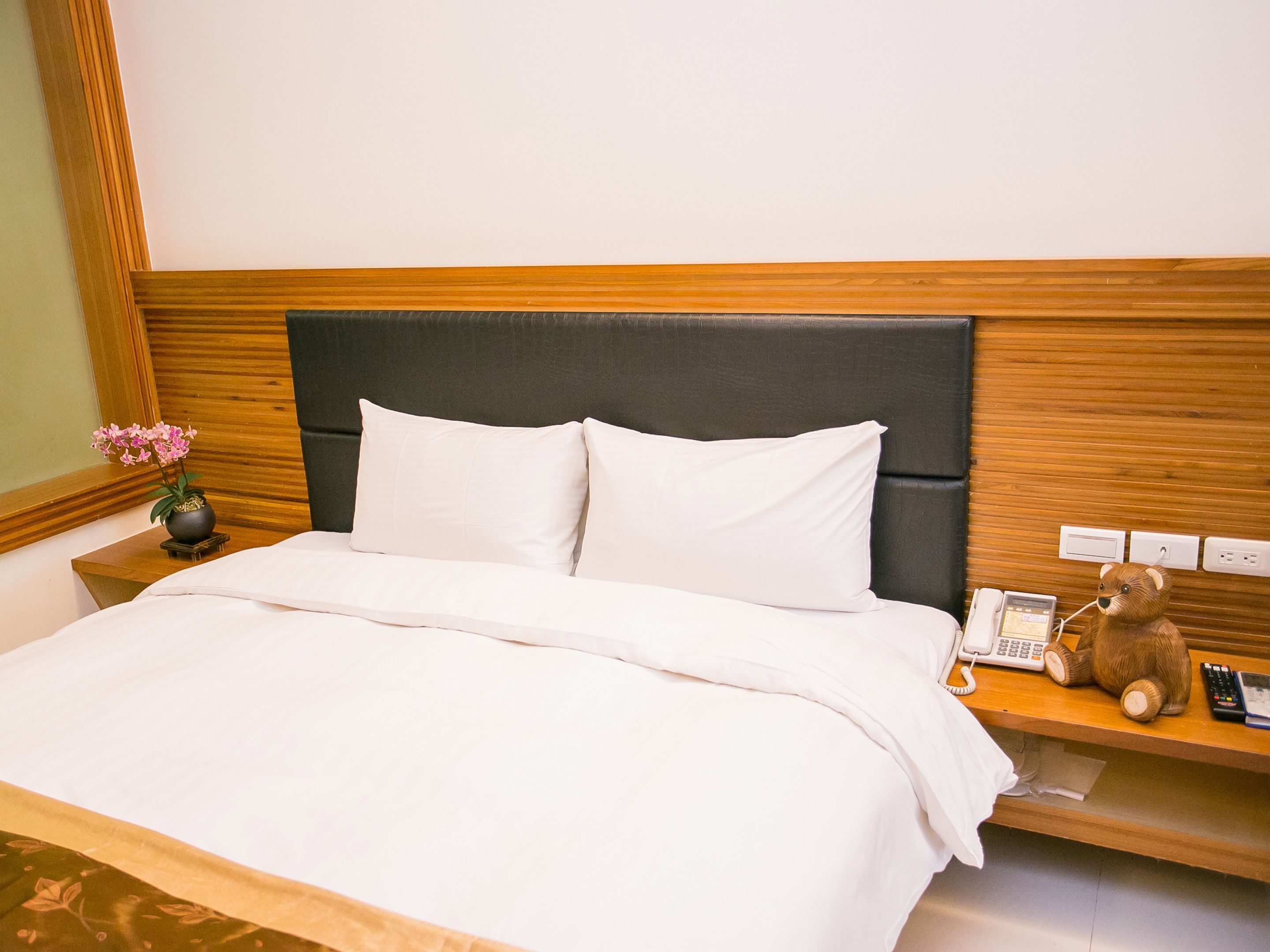 A neatly made bed with white linens, a dark headboard, and a bedside table with a teddy bear, a phone, and a vase with flowers.
