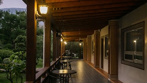 An outdoor covered patio with wooden beams and columns, featuring tables and chairs, and a view of greenery and hills in the background. - Flying Villa (Puli)