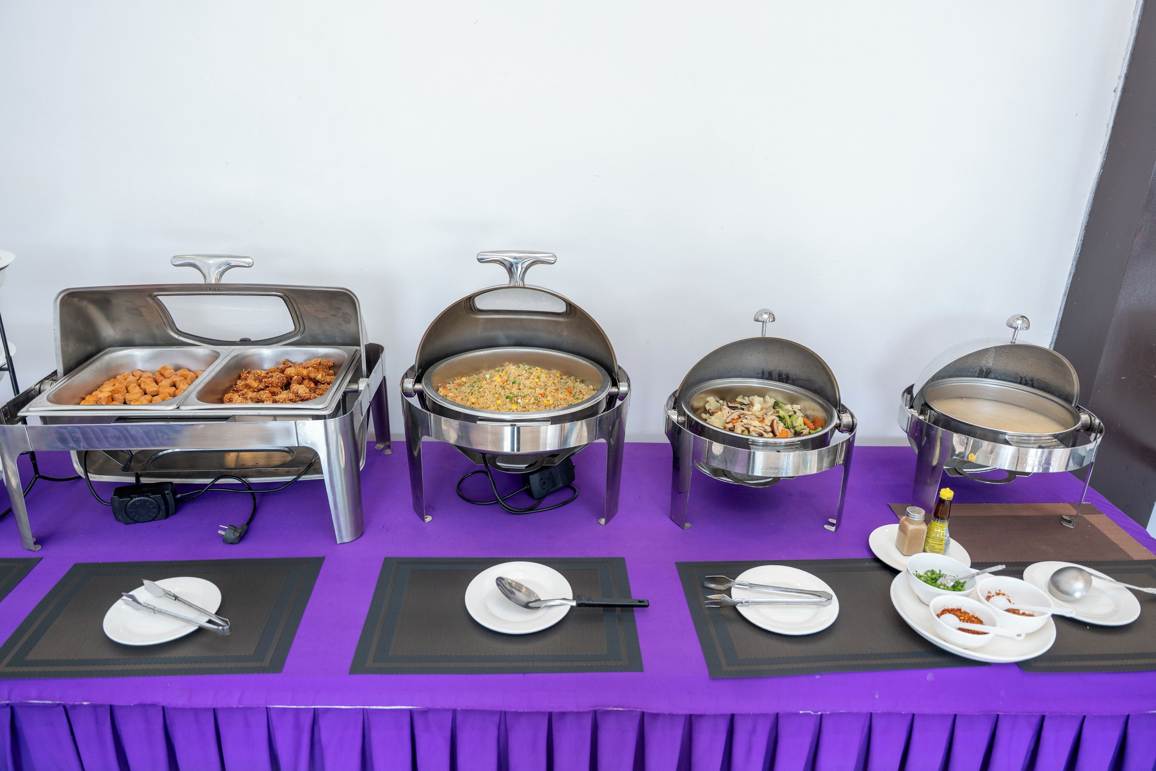 Montaje tipo bufé con varios recipientes de comida y utensilios para servir sobre un mantel morado.