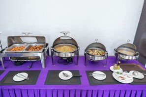 Montaje tipo bufé con varios recipientes de comida y utensilios para servir sobre un mantel morado.