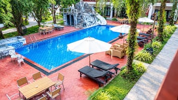 Una piscina con sillas y mesas de descanso, rodeada de follaje y una formación rocosa decorativa.