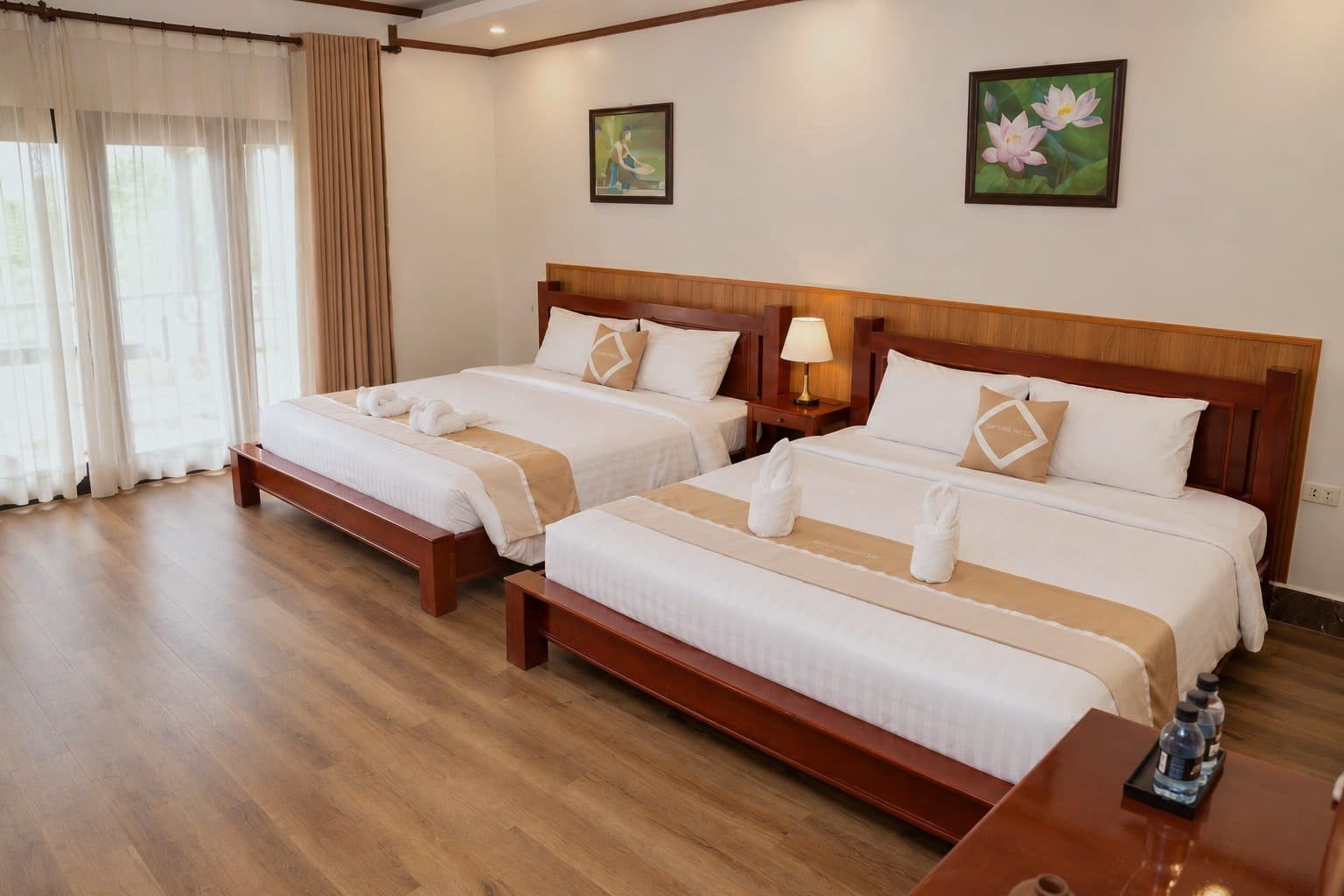 Une chambre d’hôtel avec deux lits, des têtes de lit en bois, du linge de lit blanc, une table de chevet avec une lampe et une fenêtre avec des rideaux légers.