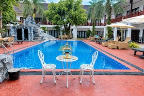 A swimming pool with white chairs and a table, surrounded by lounge chairs and a palm tree.