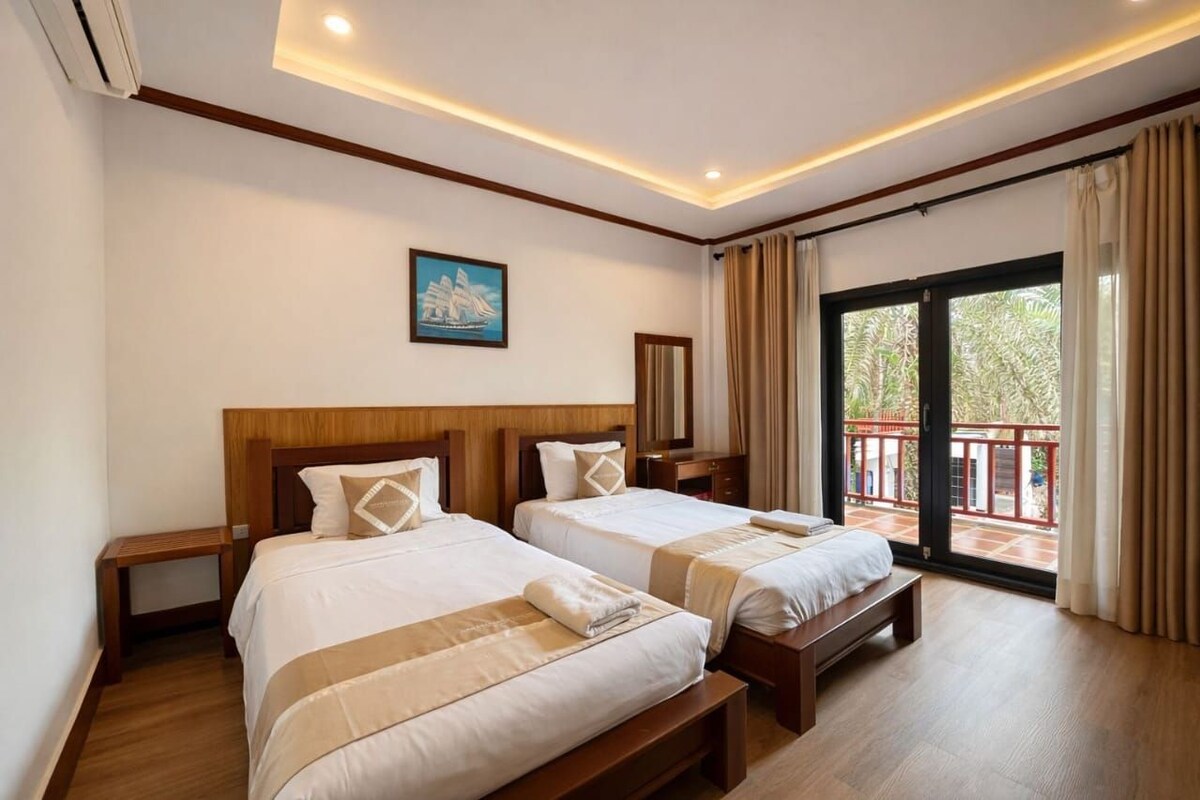 Habitación de hotel con dos camas, cabecero de madera, un cuadro en la pared y un balcón con cortinas.
