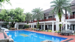 Una piscina con sillones de descanso y sombrillas, rodeada de palmeras y un edificio con balcones.