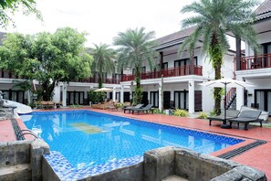Una piscina con sillones de descanso y sombrillas, rodeada de palmeras y un edificio con balcones.