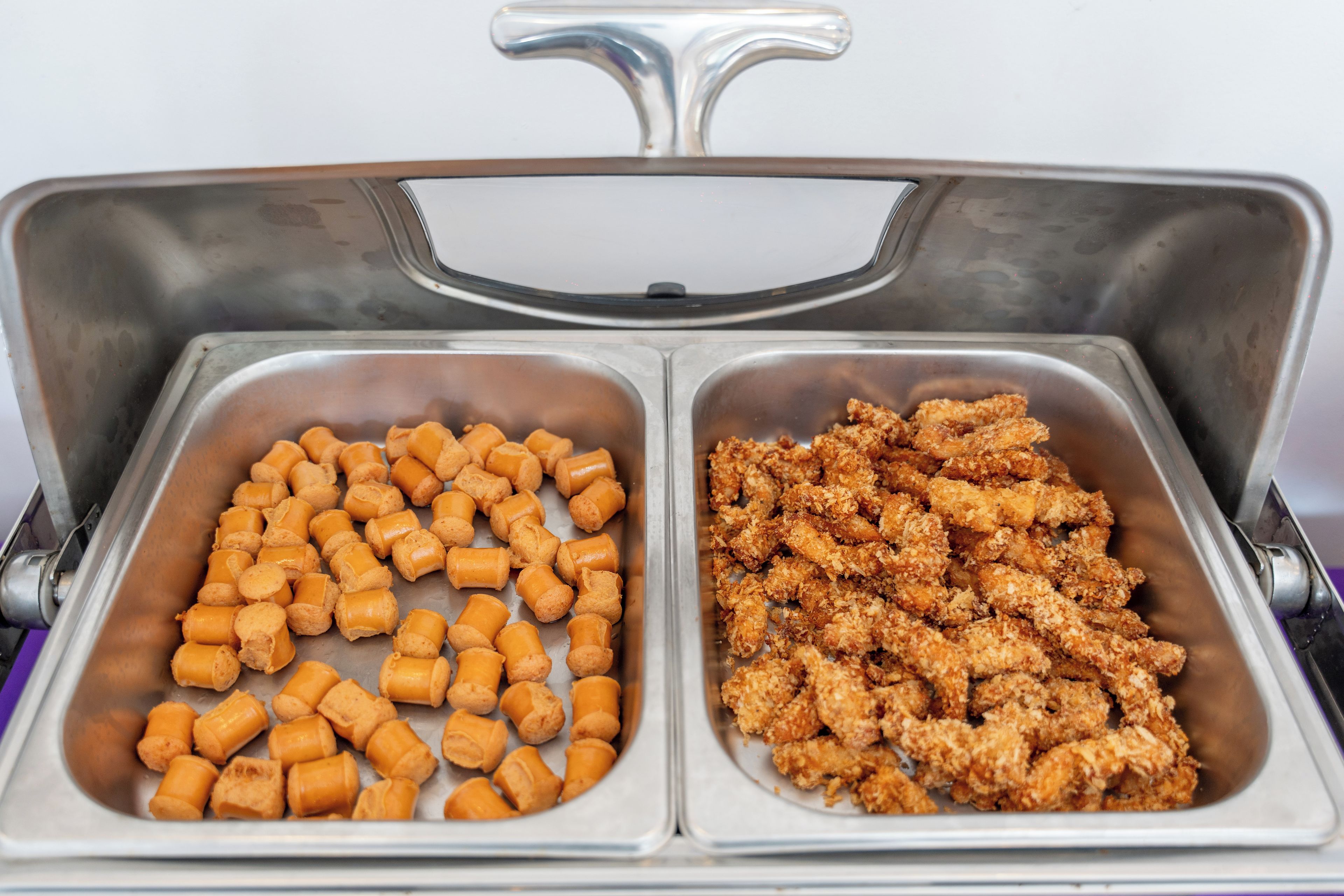 A stainless steel buffet with two compartments, one containing cylindrical food items and the other containing breaded chicken pieces.
