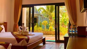 A hotel room with a bed, a small table, a TV, and a sliding glass door leading to a garden.