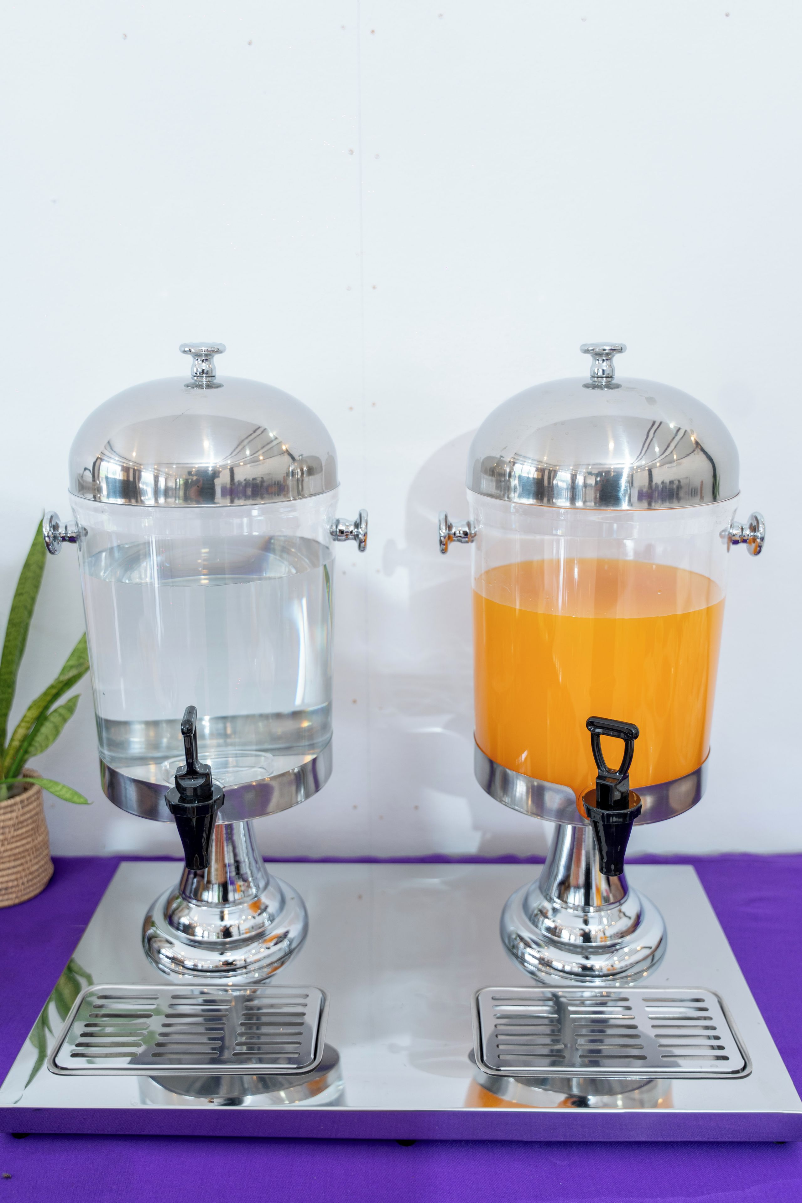 Two beverage dispensers with transparent tanks and metallic tops, mounted on a tray with a reflective surface.