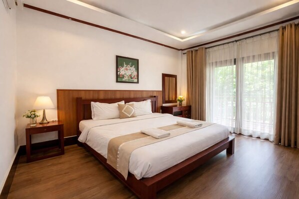 Habitación de hotel con una cama grande, cabecera de madera, mesitas de noche con lámparas, un cuadro en la pared y una ventana con cortinas.