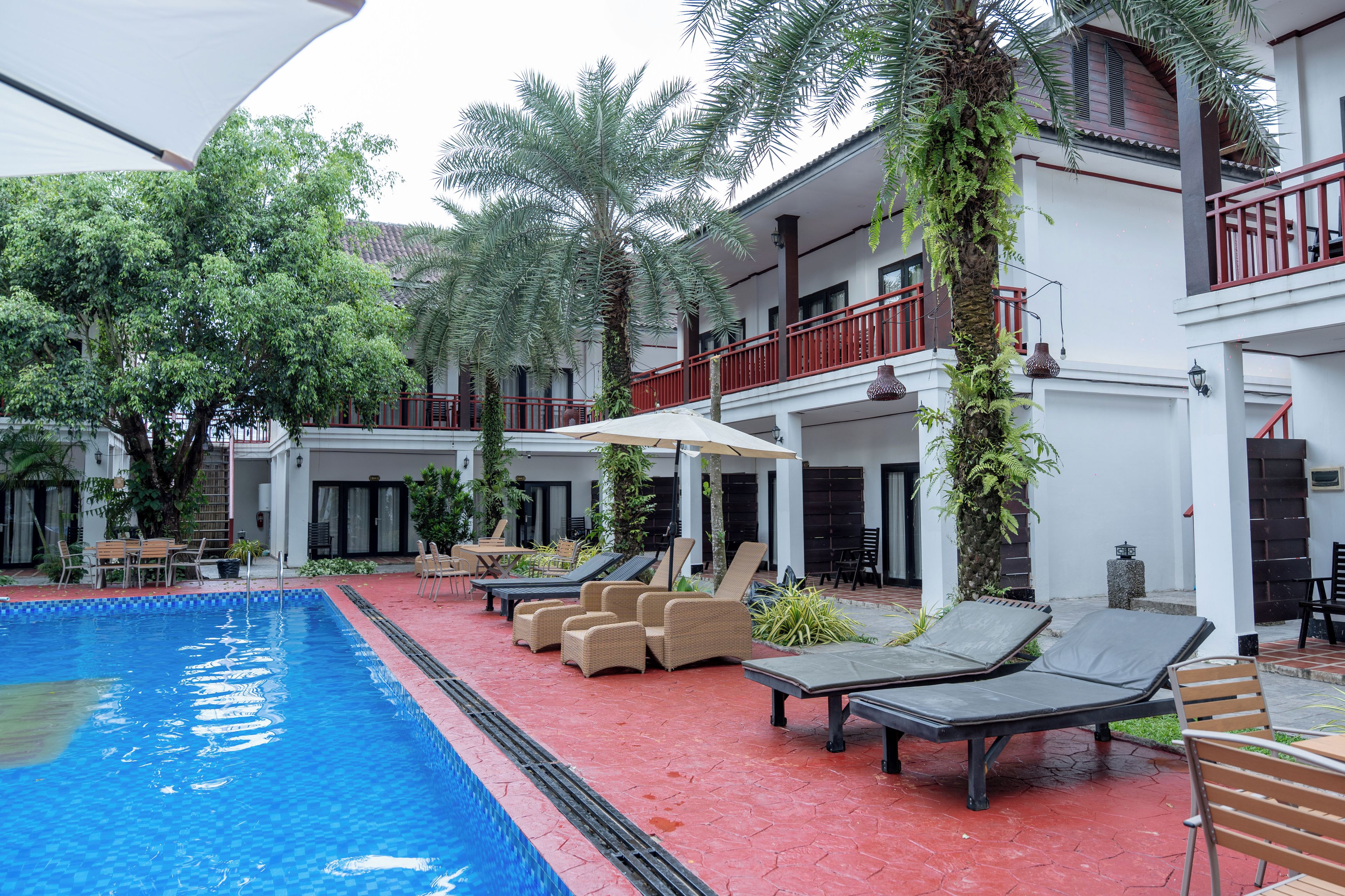 Un espace aménagé au bord de la piscine, avec des chaises longues et des parasols, entouré de verdure et bordé d’un bâtiment doté d’un balcon avec une rambarde rouge.