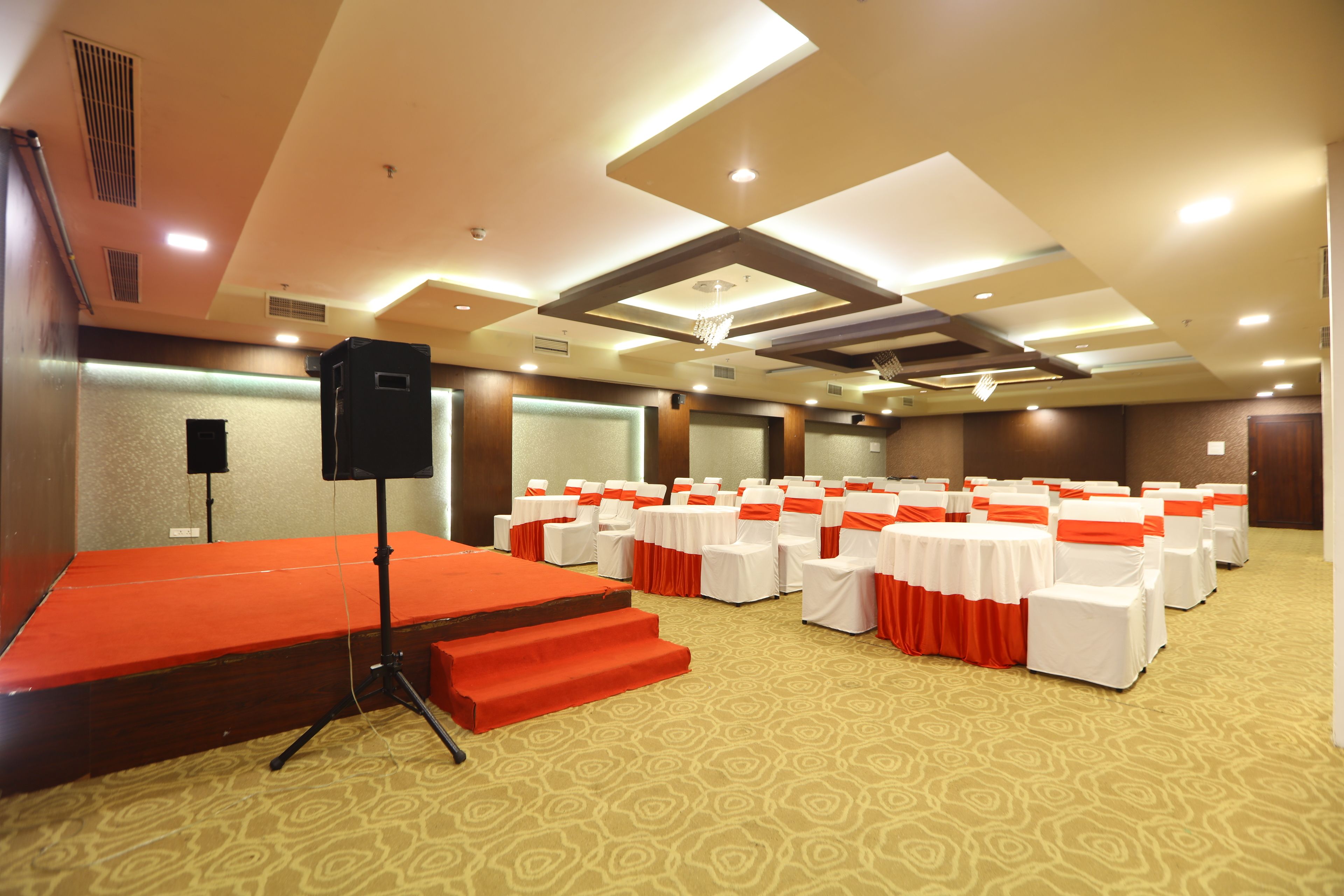 A conference room with a stage, rows of tables with white tablecloths and orange sashes, and a carpeted floor.