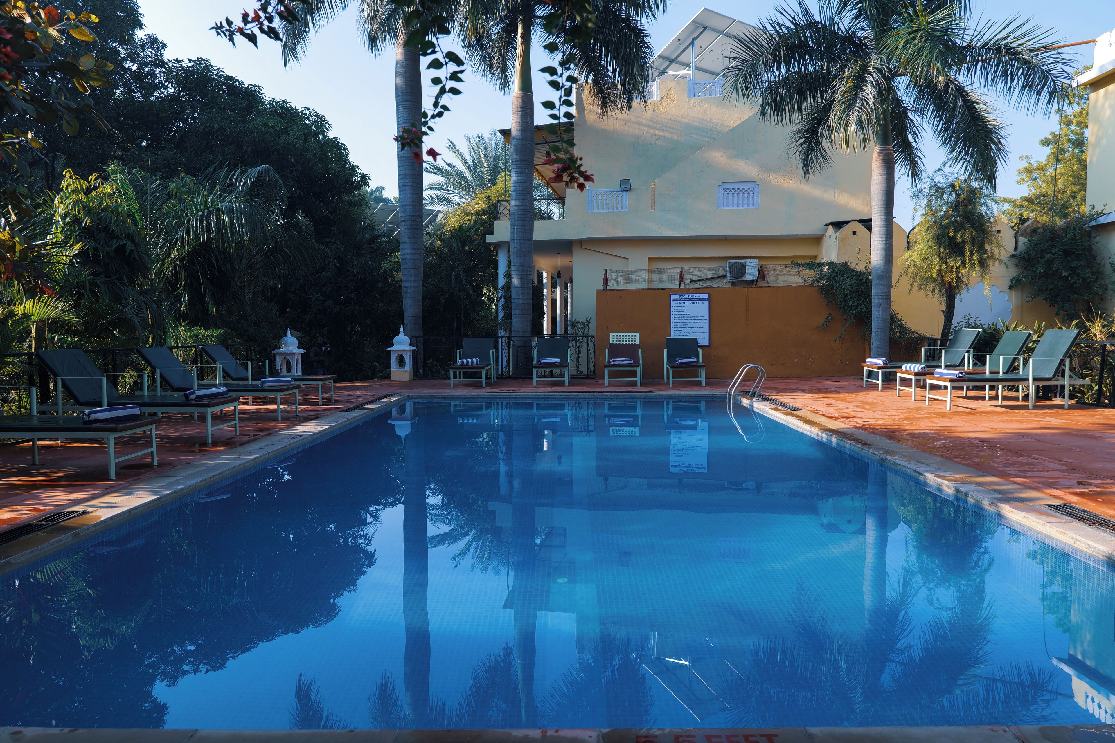 A swimming pool with lounge chairs and a small pavilion, surrounded by palm trees and a building with a balcony.