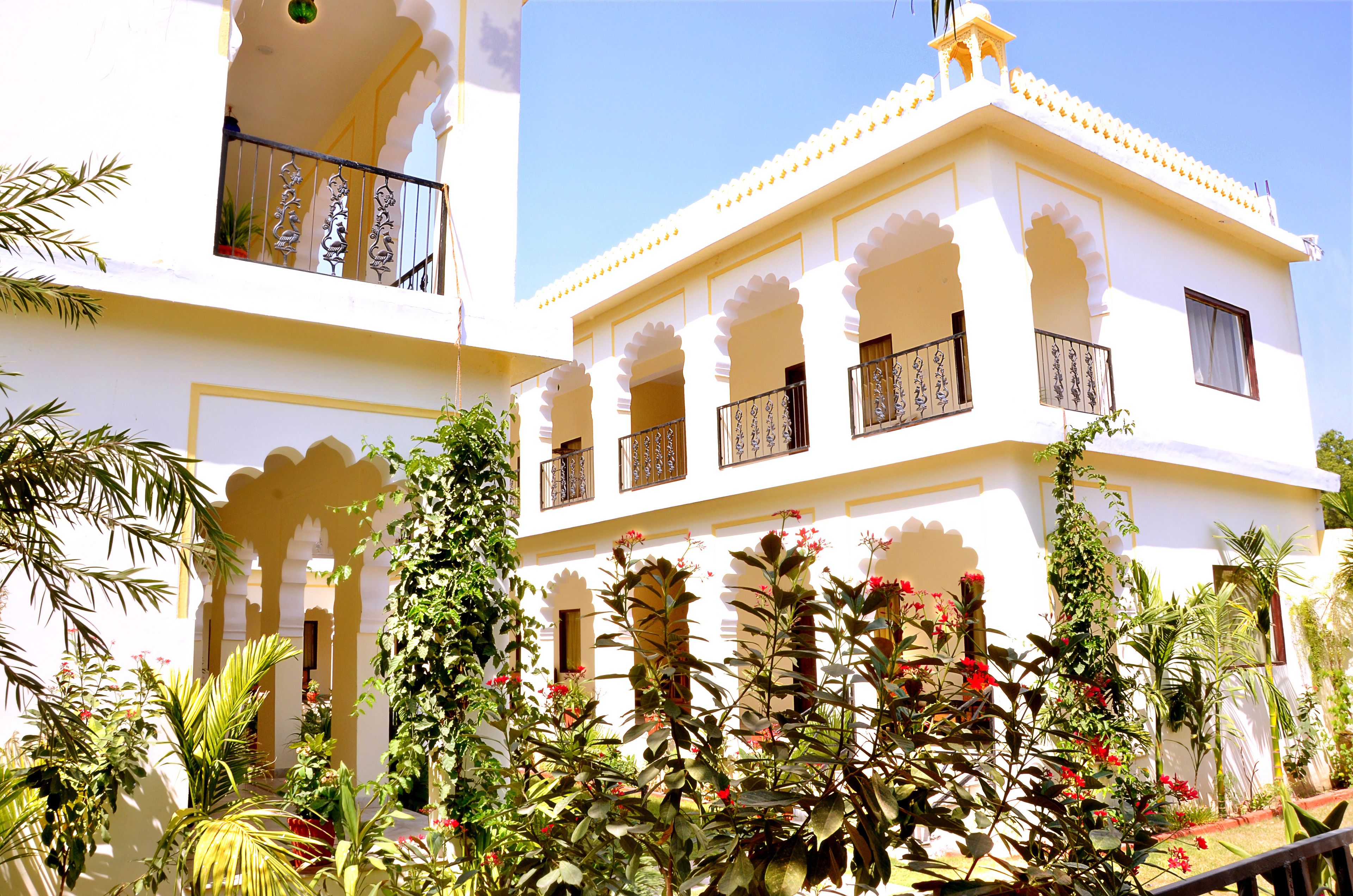 A multi-story building with balconies and arches, surrounded by lush greenery and flowering plants.