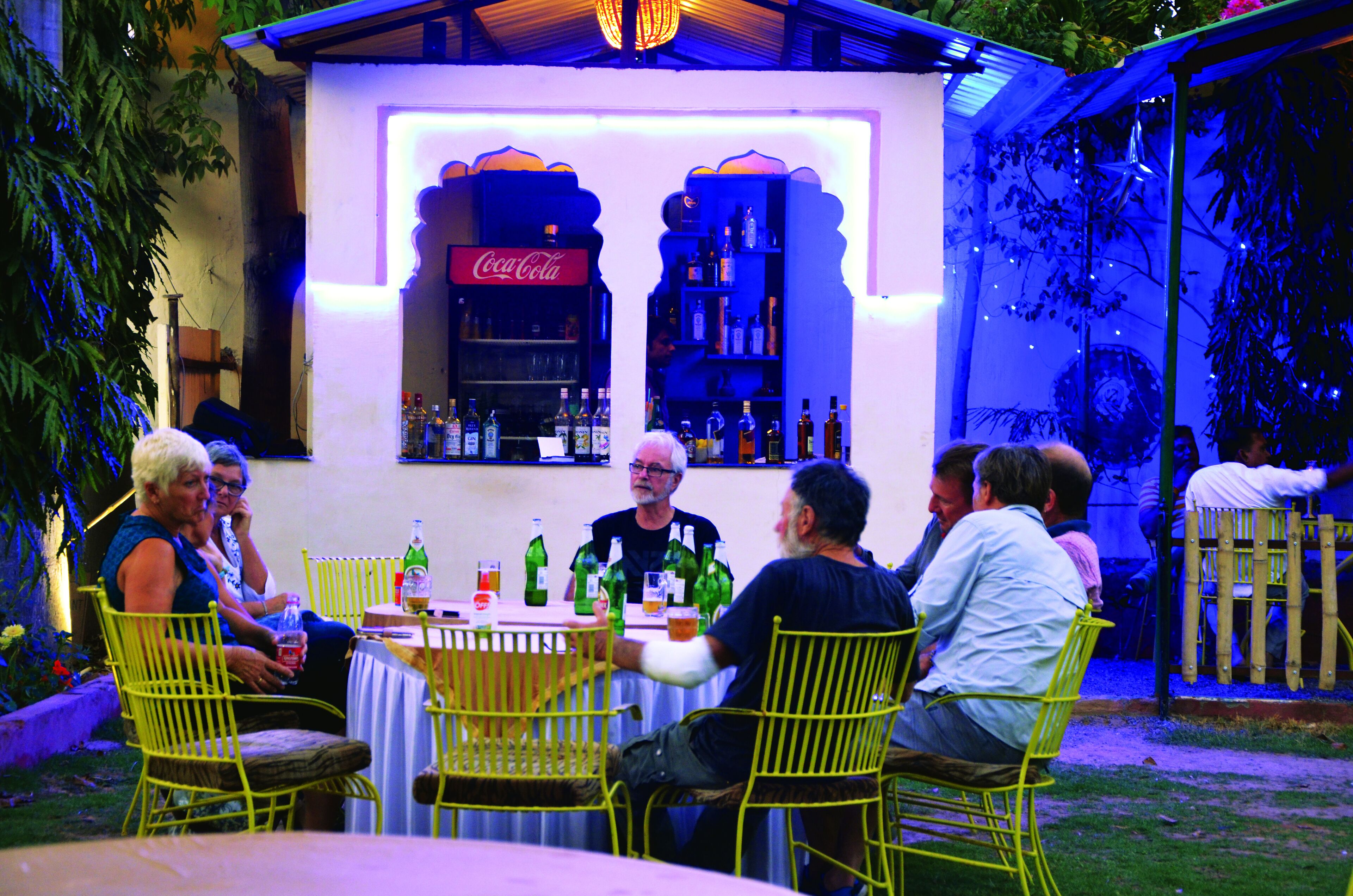 Un grupo de personas sentadas alrededor de una mesa, con un letrero de Coca-Cola arriba, en un espacio al aire libre con sillas amarillas y un mantel blanco.