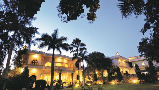 A large, well-lit mansion with multiple levels and balconies, surrounded by lush greenery and palm trees. - Raj Palace Resort (Sawai Madhopur)