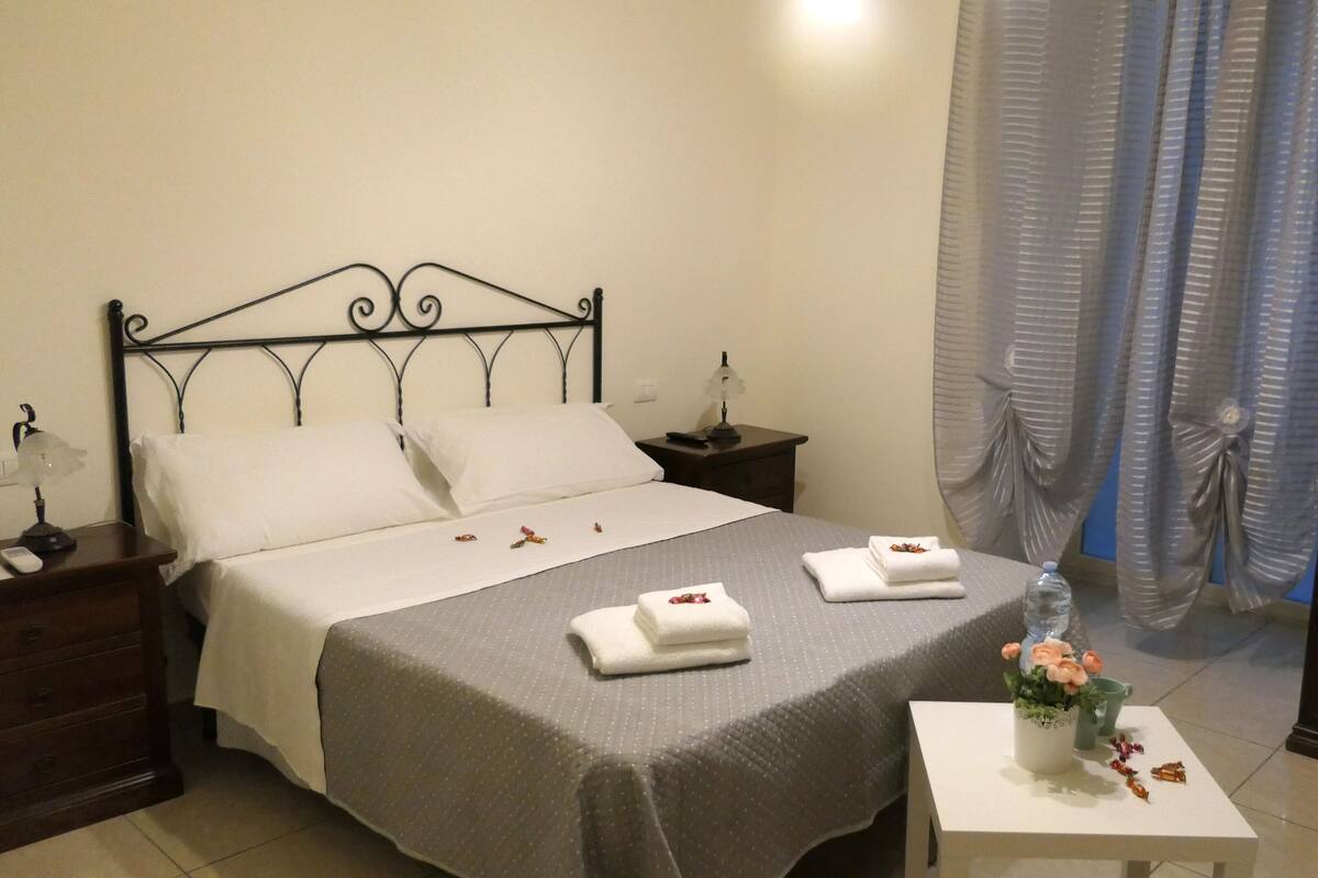 A neatly made bed with pillows, a bedside table with a lamp, a small table with a vase of flowers, and a wardrobe.