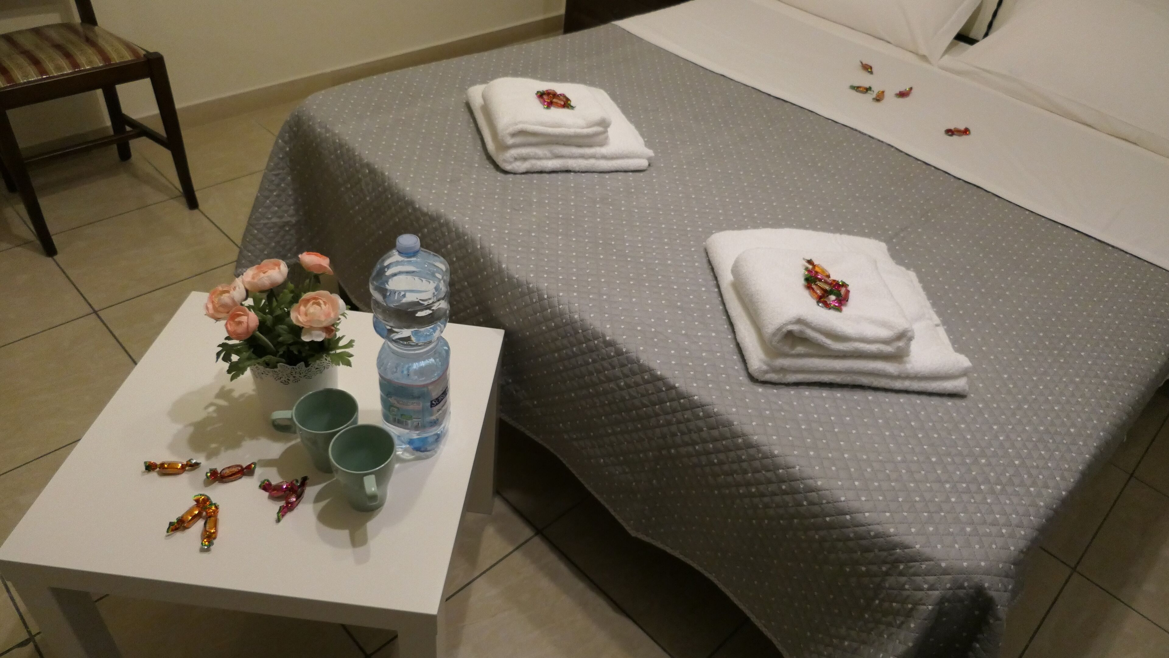 A neatly made bed with a grey quilt, two folded white towels with floral decorations, a water bottle, two green mugs, and a small table with a potted plant and decorative items.