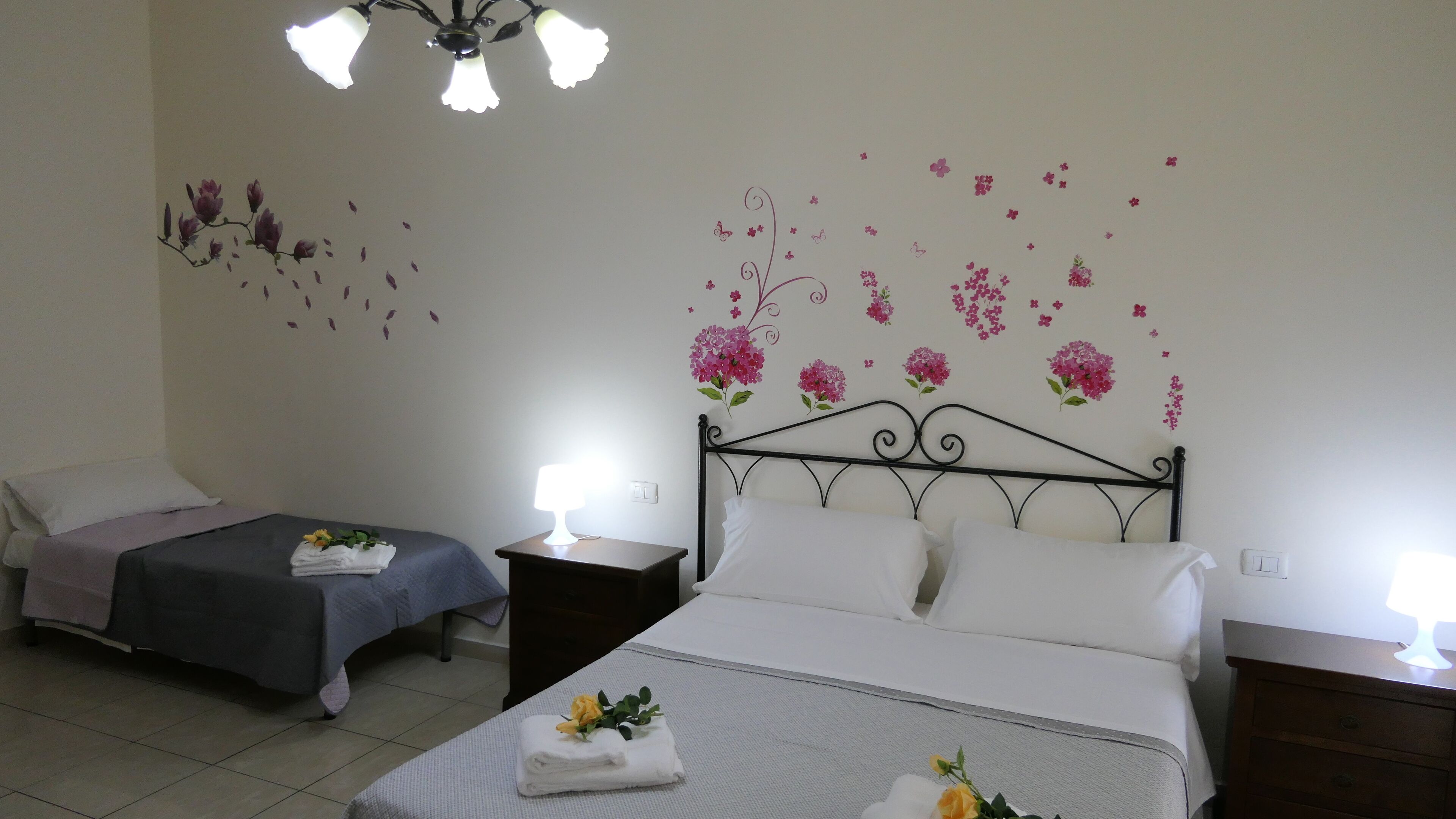 A hotel room with two beds, each with a bedside table and lamp. The room has floral wall decals and a chandelier.