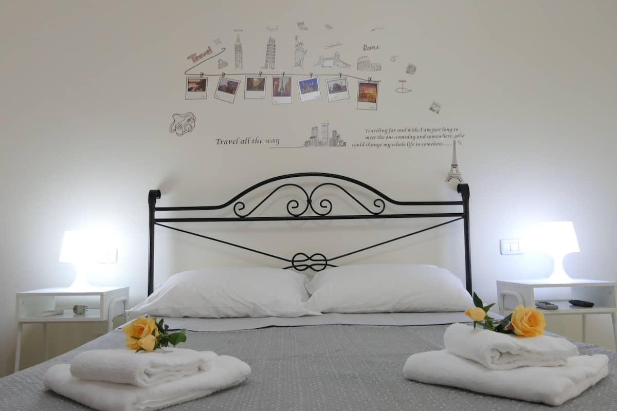 A neatly made bed with white linens and two towels, a wooden chair, and a wall with travel-themed drawings and text.