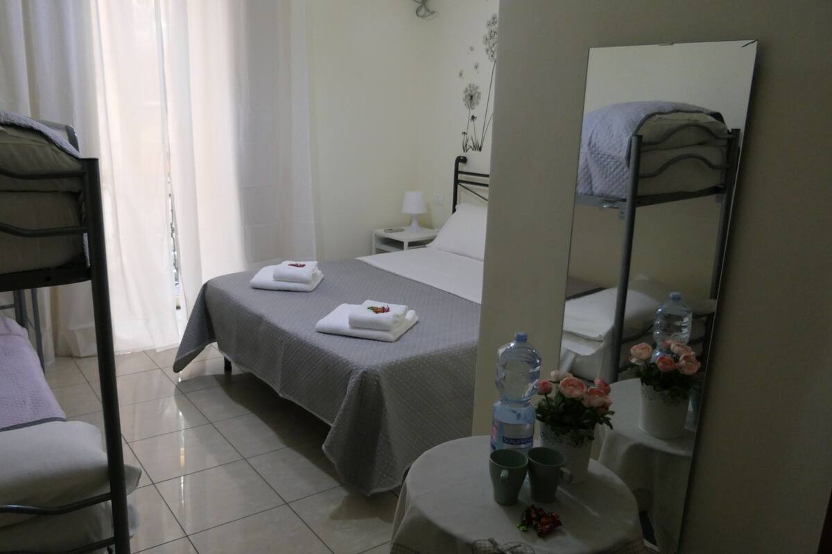 A hotel room with a bunk bed, a single bed with a grey bedspread, a bedside table with a lamp, a mirror, a small table with a water bottle and flowers, and a window with curtains.