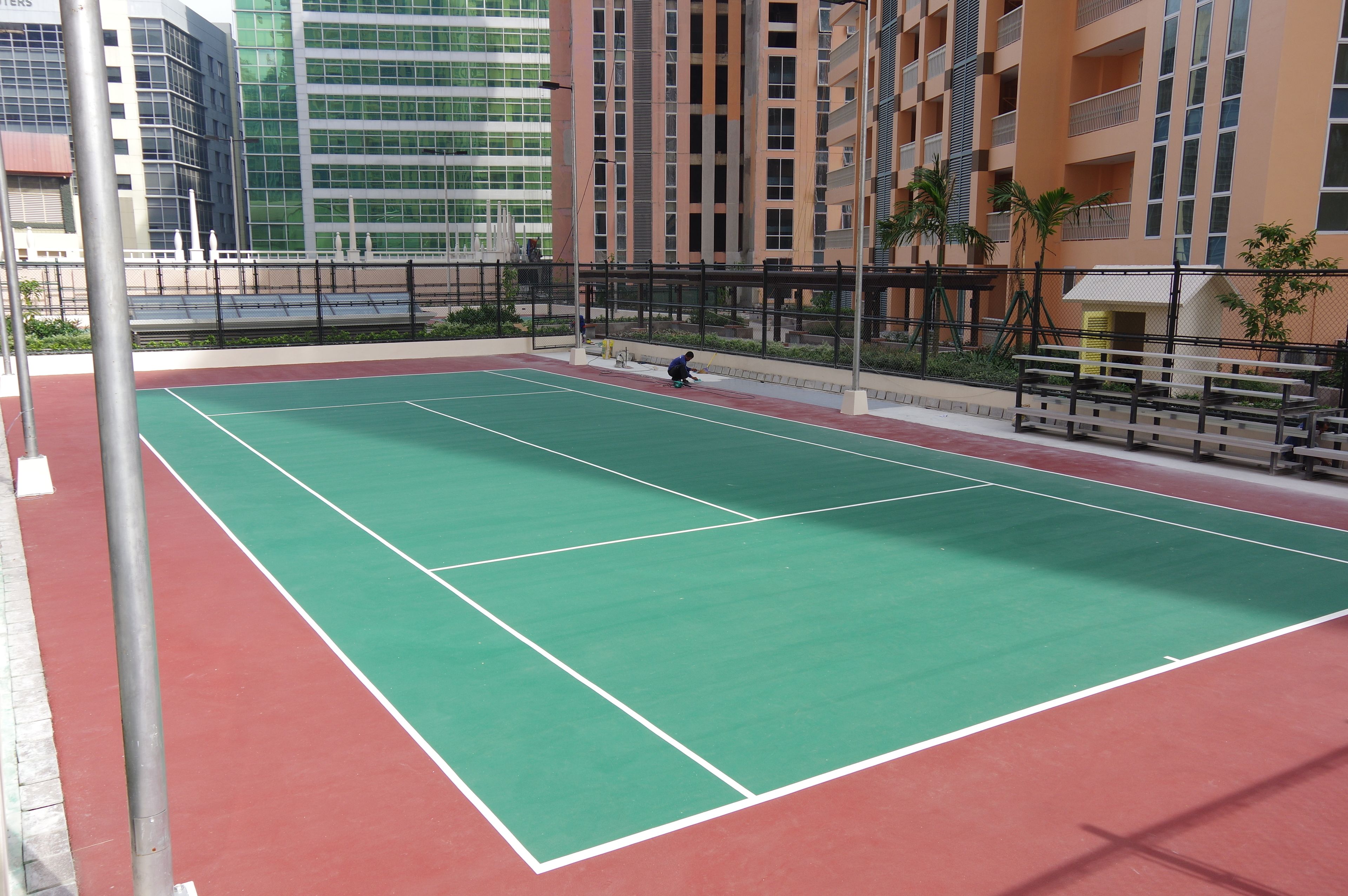 Una cancha de tenis al aire libre con superficie roja y verde, rodeada por una valla y edificios al fondo.