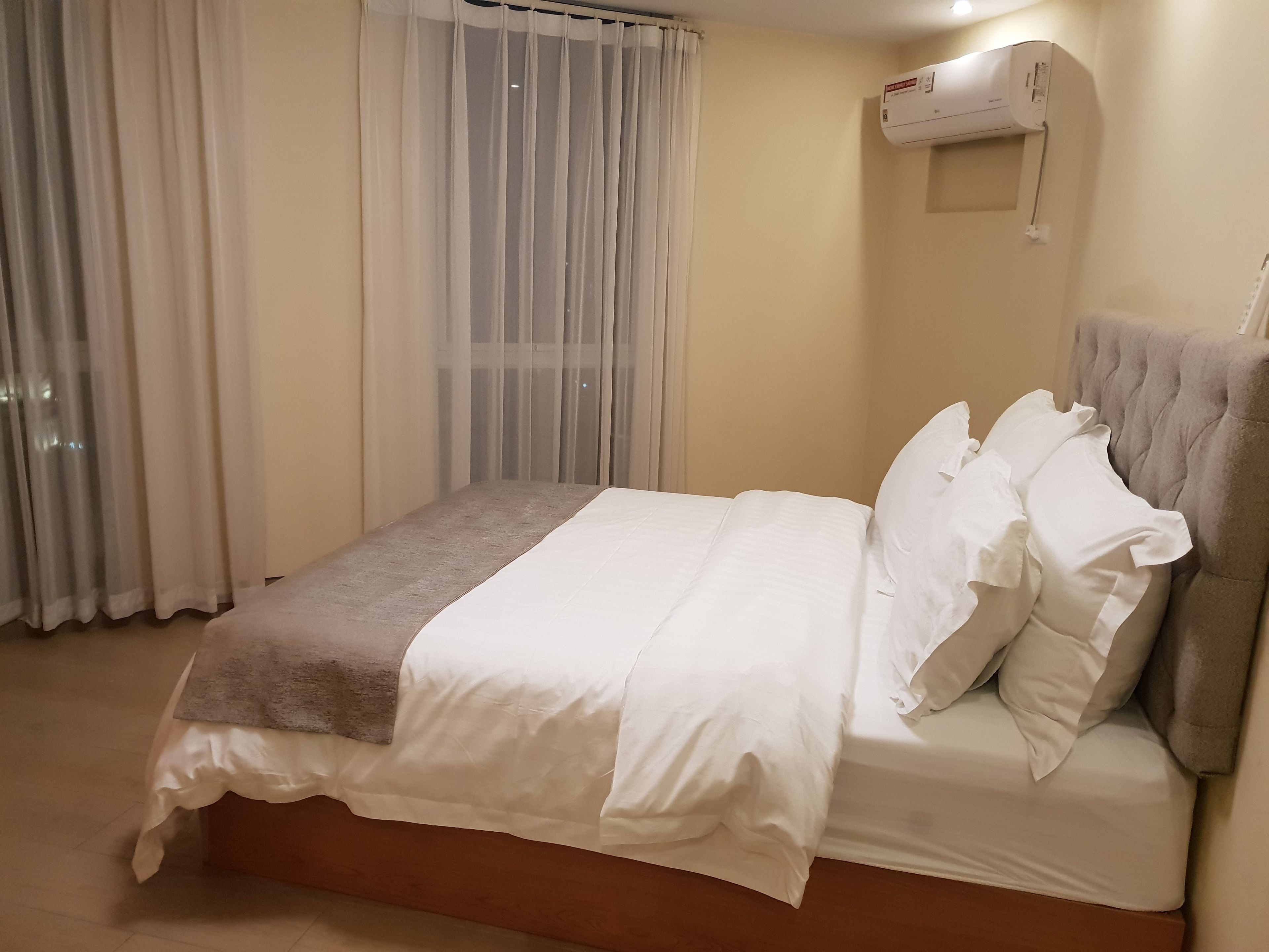 A neatly made bed with white linens and a brown throw, a grey upholstered headboard, and a wall-mounted air conditioning unit.