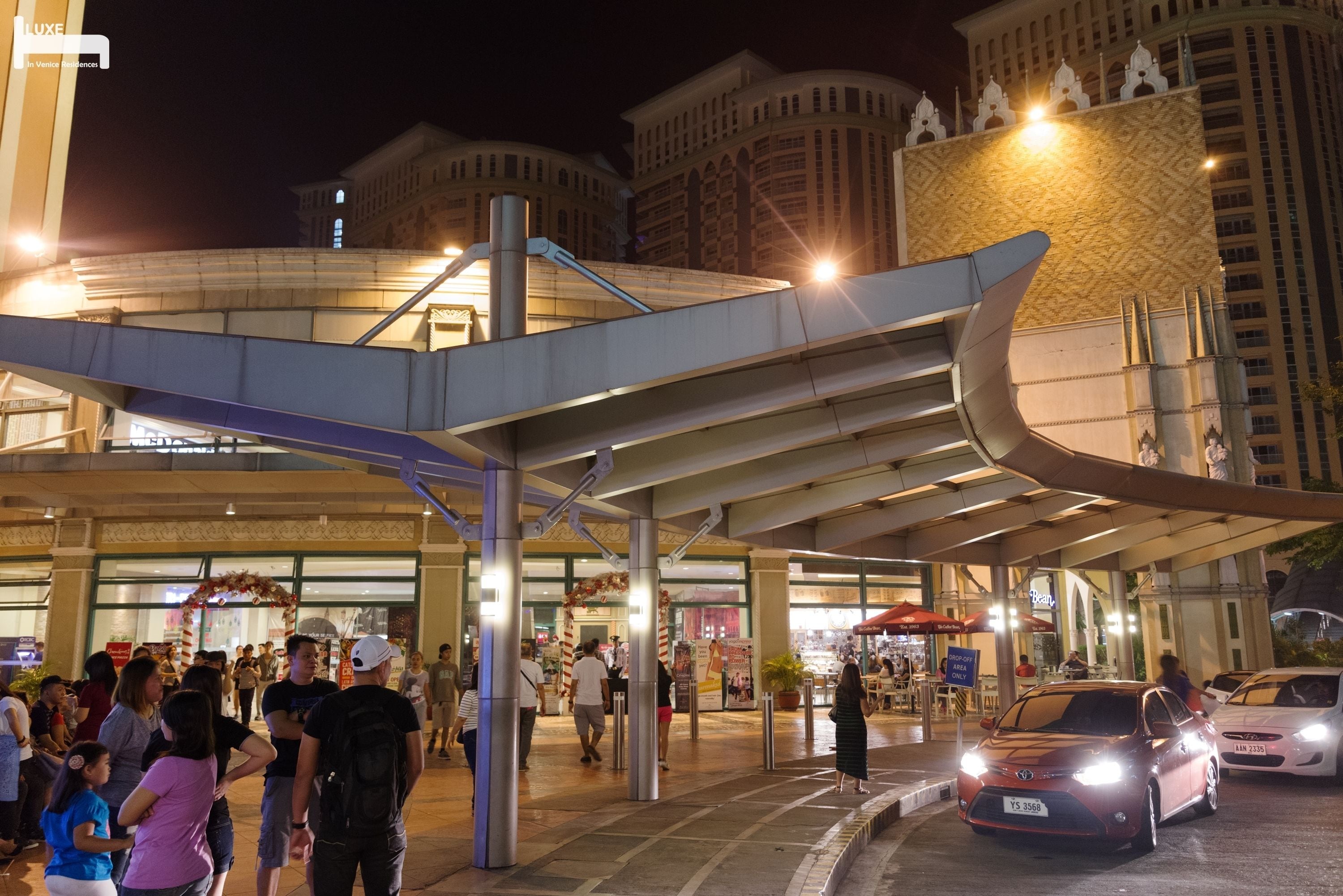 A hotel entrance with a covered walkway, a crowd of people, and a parking area with cars.