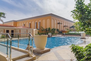 Zona de piscina con un edificio grande al fondo, una planta en maceta grande y una persona en el agua.