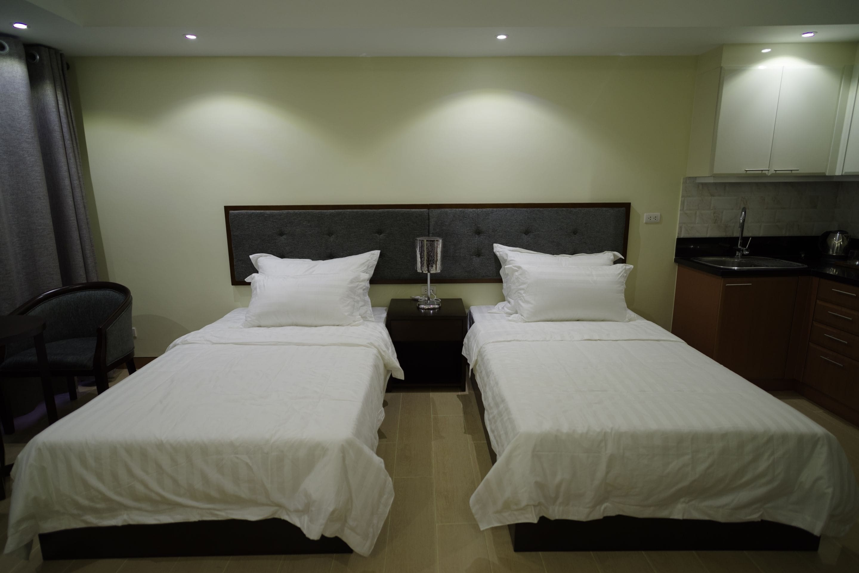 A hotel room with two single beds, a nightstand, a chair, and a kitchenette.