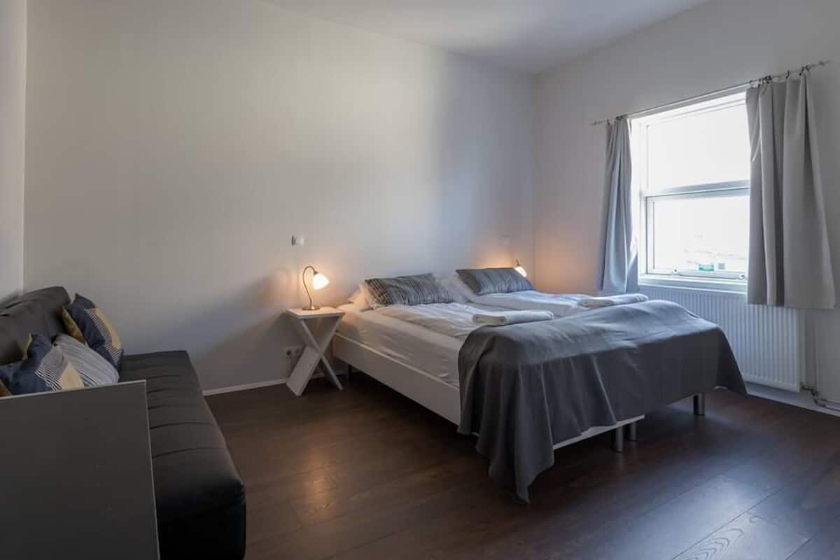 Un dormitorio con cama, mesitas de noche, una ventana con cortinas y un sofá.