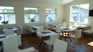 A spacious room with multiple white tables and chairs, large windows with a view of buildings and hills, and a wooden floor. - Hotel Eyjar (Vestmannaeyjar)