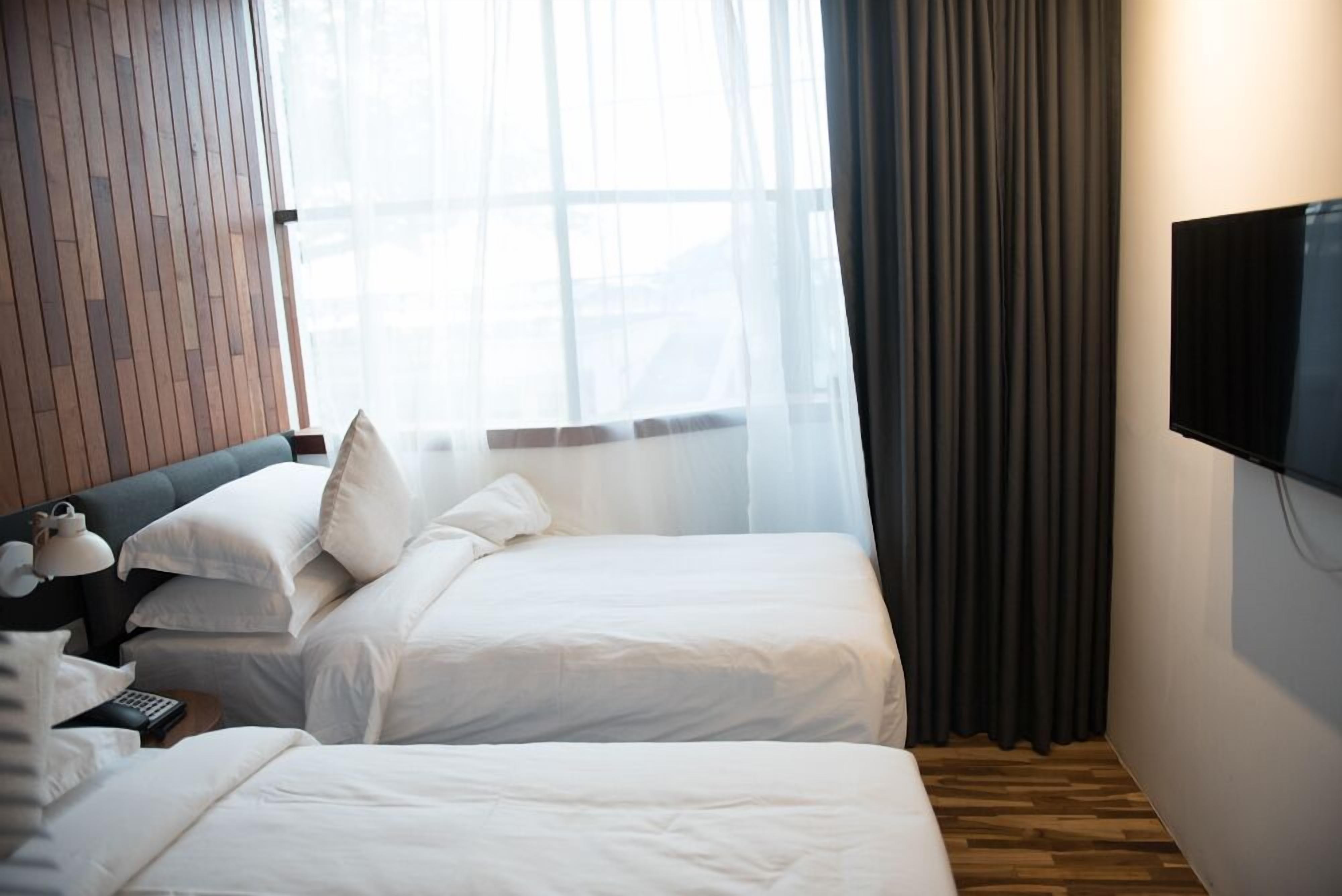 A hotel room with two beds, a wooden headboard, a large window with sheer curtains, a flat-screen TV mounted on the wall, and a wooden floor.