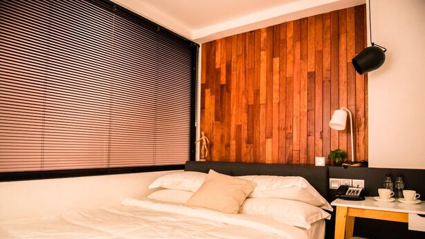 Un dormitorio moderno con pared de paneles de madera, una cama con ropa de cama blanca, un escritorio con una silla, y una mesita de noche con una lámpara y un teléfono.