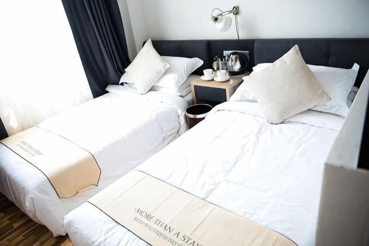A hotel room with two beds, a nightstand with a lamp, and a view of the outdoors.