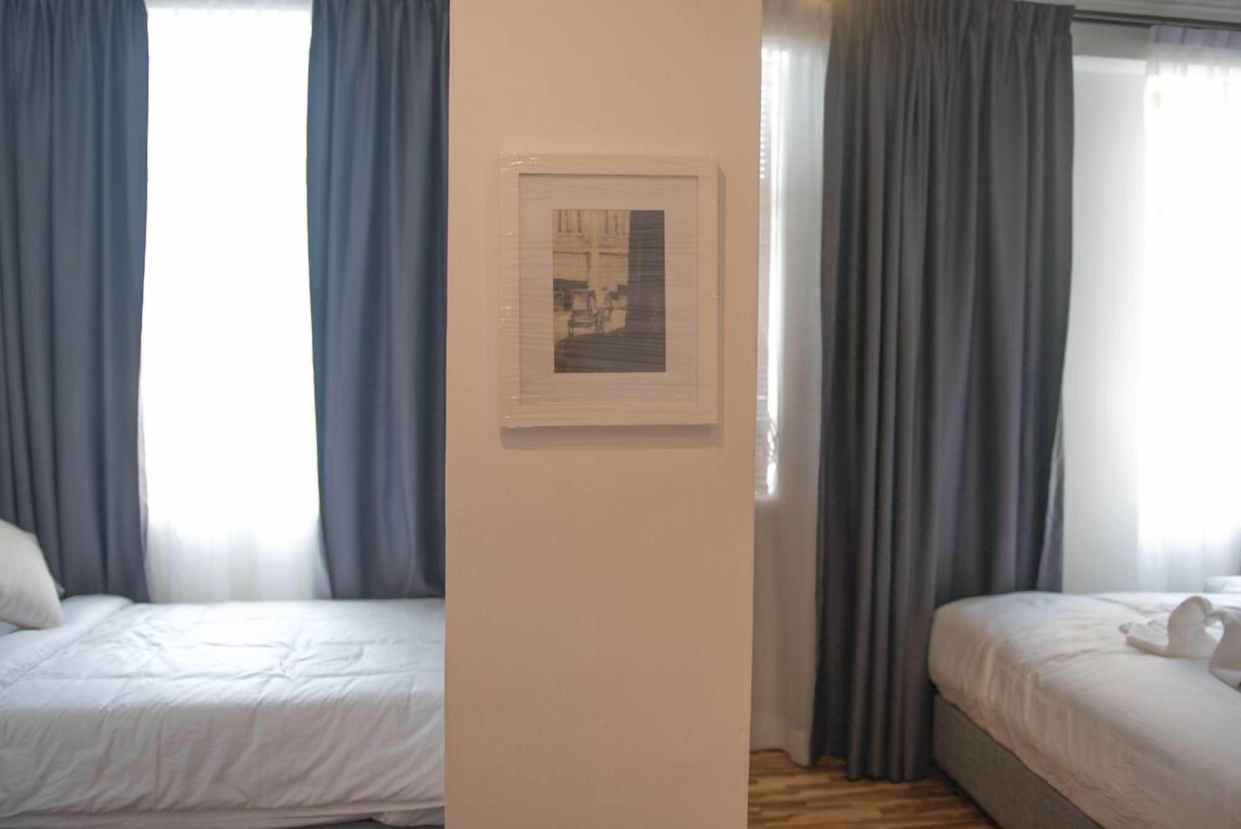 Quarto de hotel com duas camas, um quadro na parede e cortinas cinzentas.
