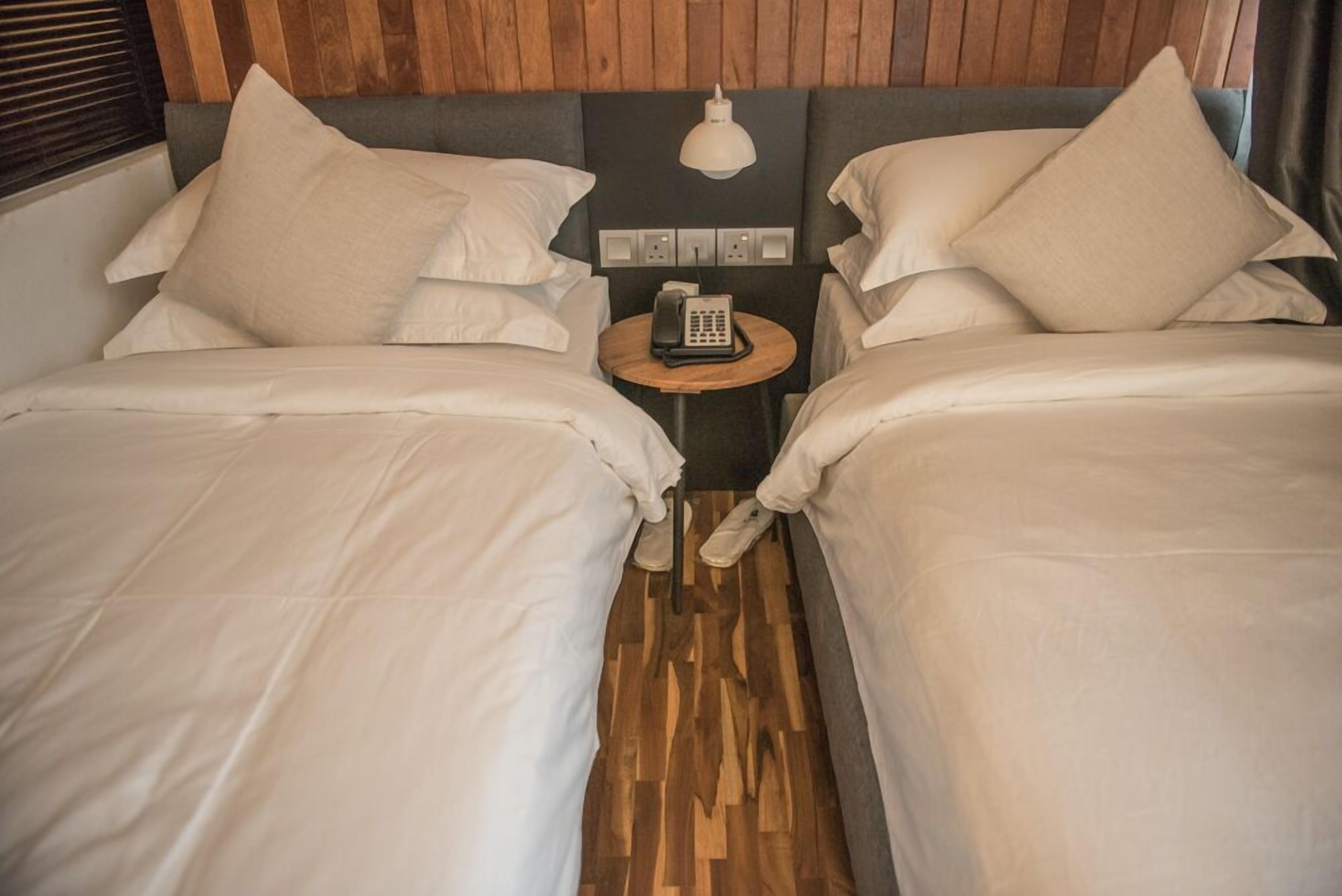 A hotel room with two beds, a wooden floor, a small table with a phone, and a wall-mounted light fixture.